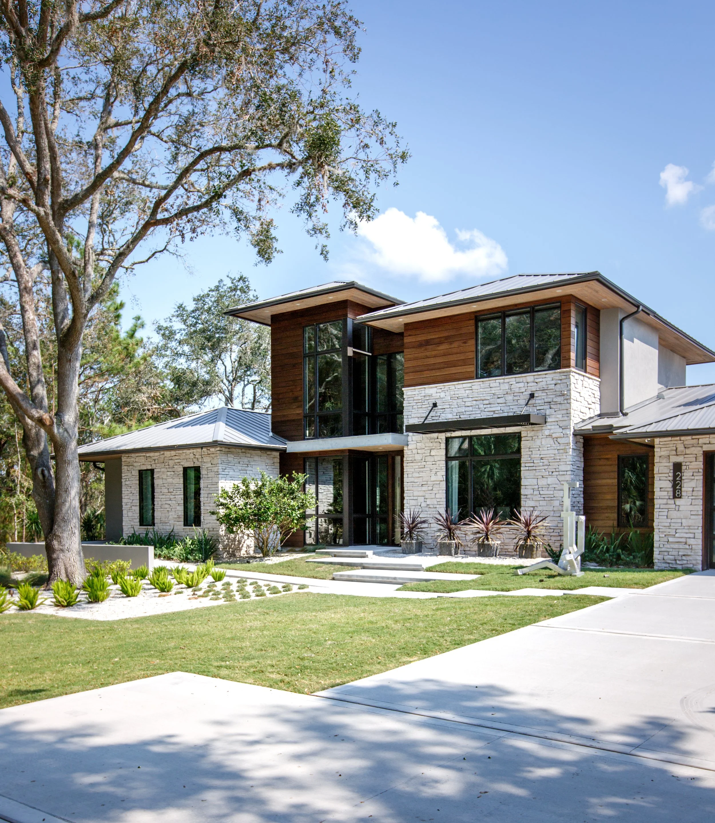 Modern two-story house with large windows, wooden and stone exterior, front yard with green grass, small shrubs, and a sidewalk. Tall trees and a blue sky with clouds are in the background.