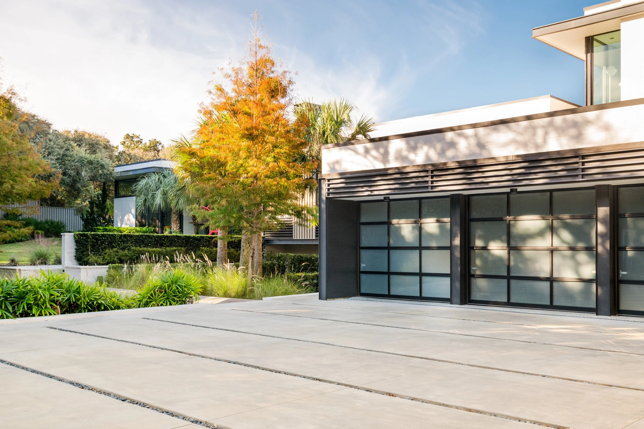 Modern house with large glass windows, surrounded by greenery including palm and deciduous trees with fall foliage, on a clear day.