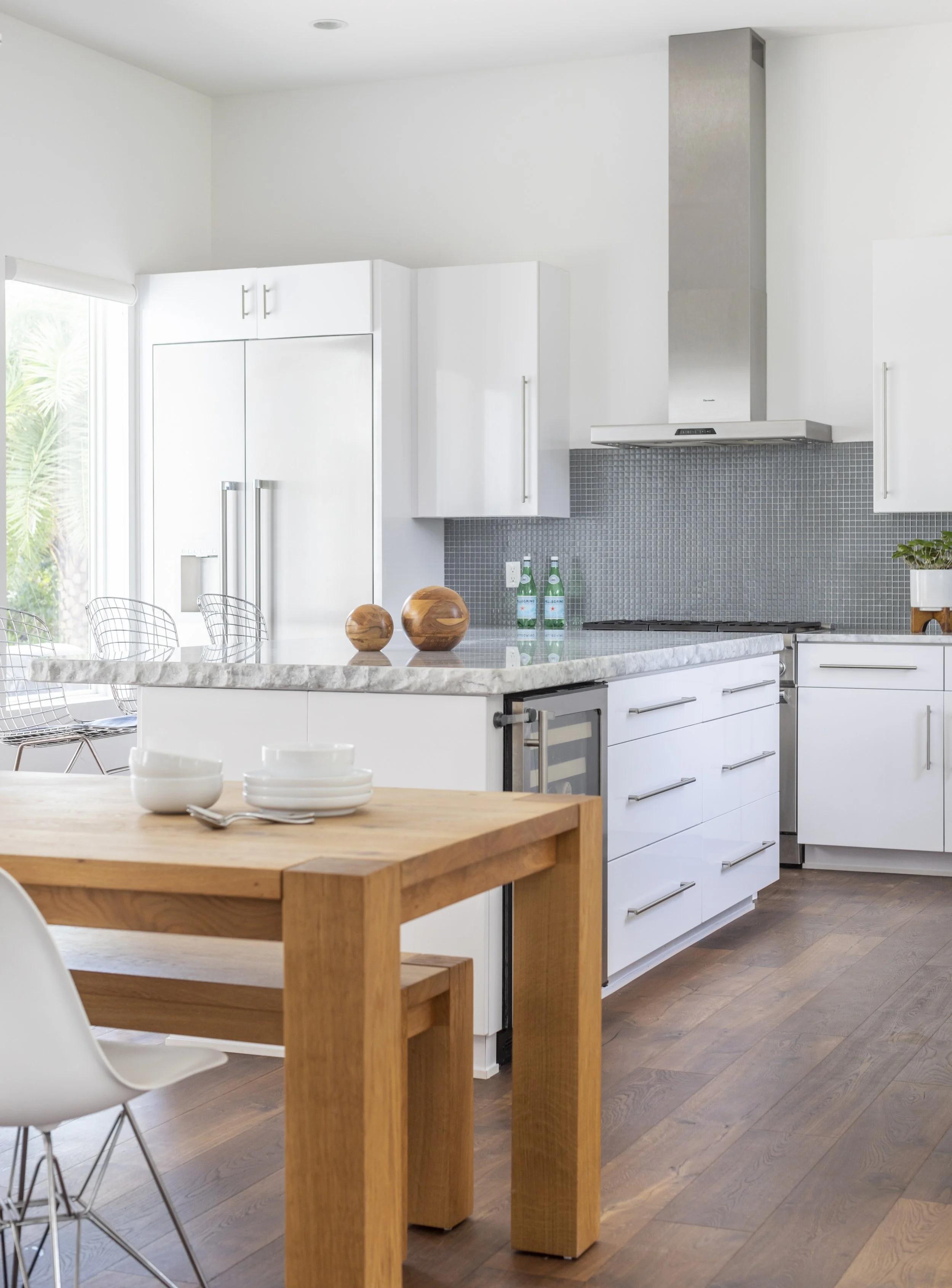 A modern kitchen with white cabinets, a marble island, a wooden dining table, metal wire chairs, a grey tiled backsplash, and a stainless steel range hood. Two bottles of sparkling water and wooden decorative objects are on the marble island.