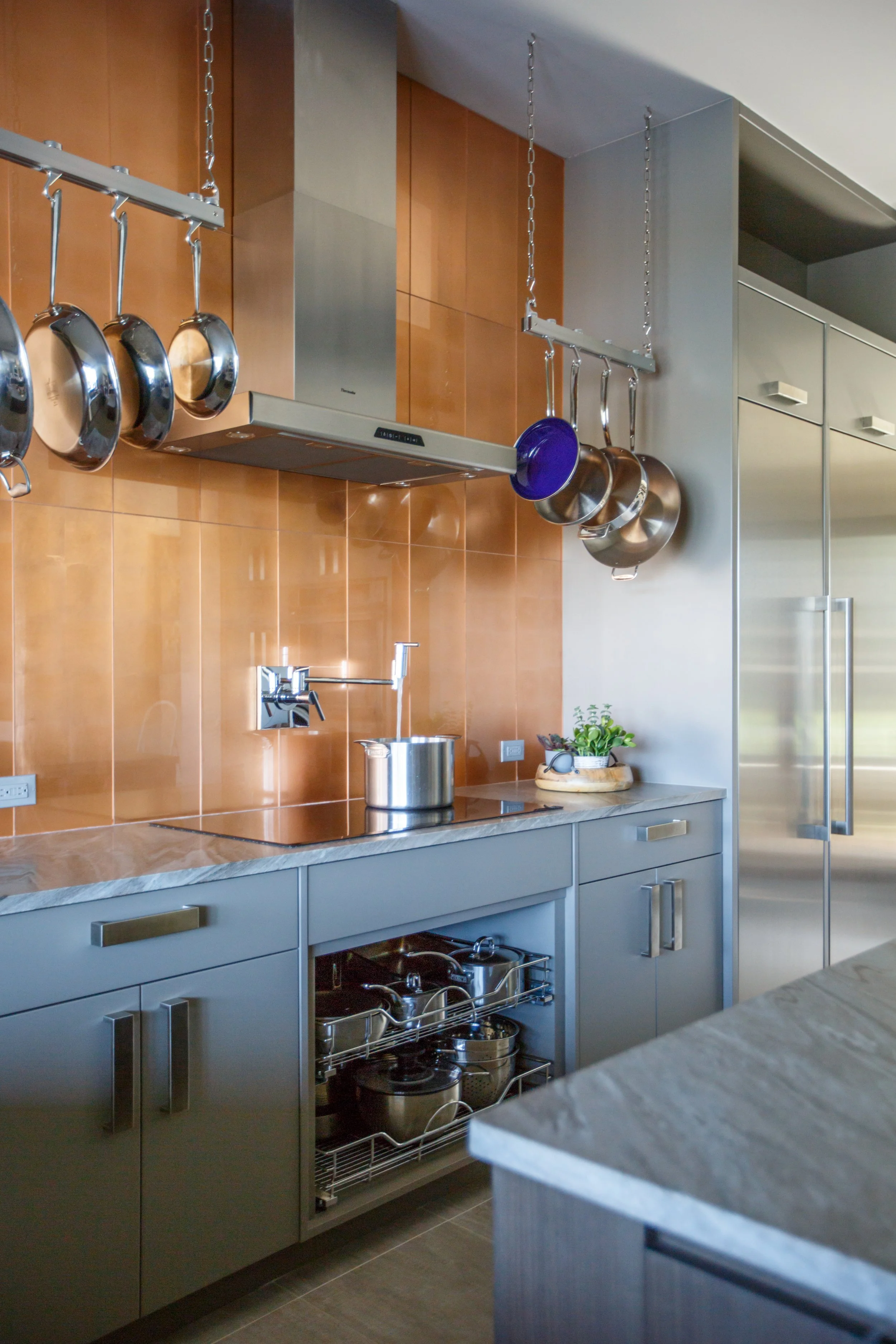 Modern kitchen with gray cabinets, a marble countertop, a stainless steel exhaust hood, and hanging pots and pans.