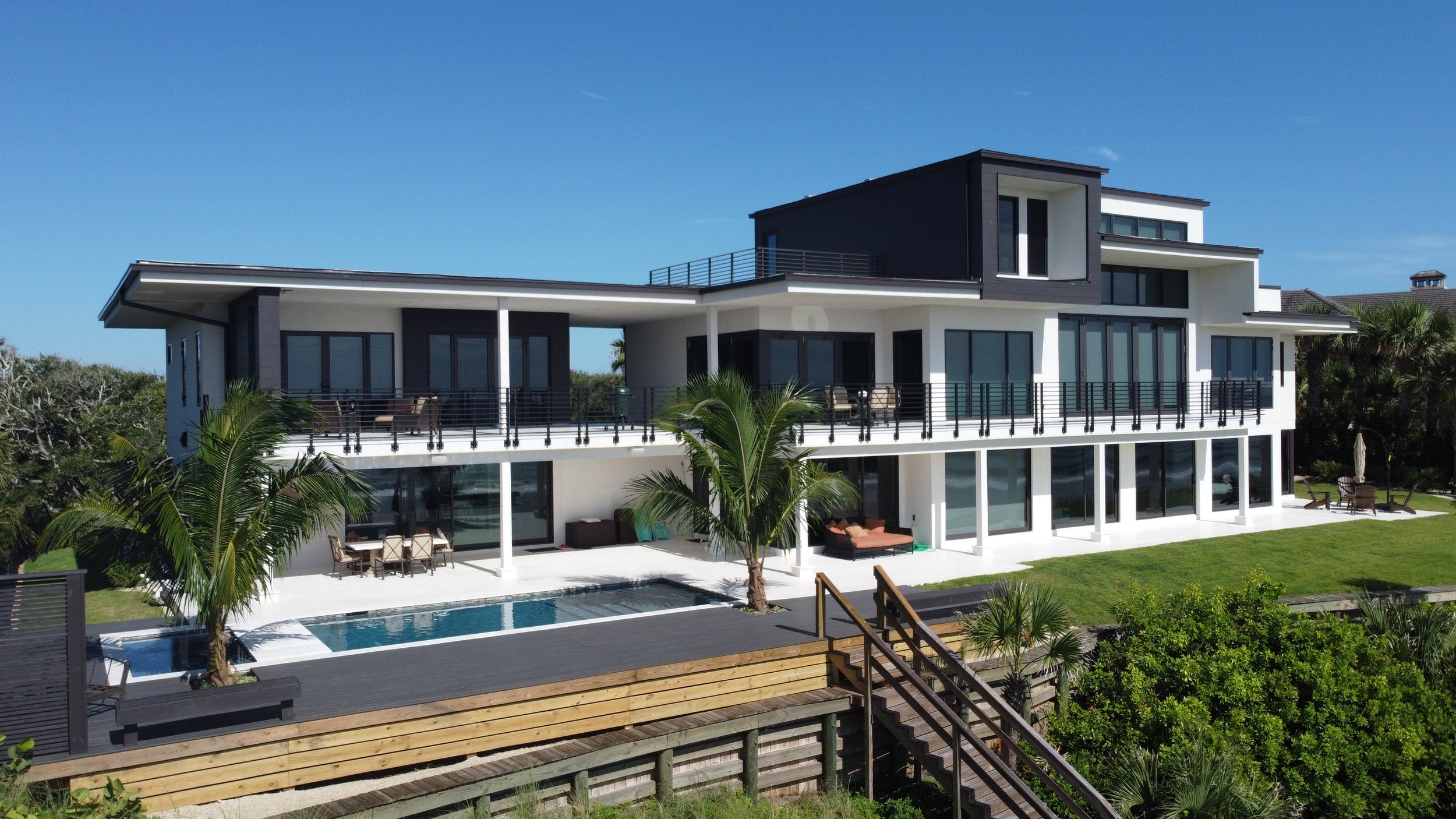 Modern multi-story house with large glass windows, balconies, surrounded by palm trees and a backyard pool under a clear blue sky.