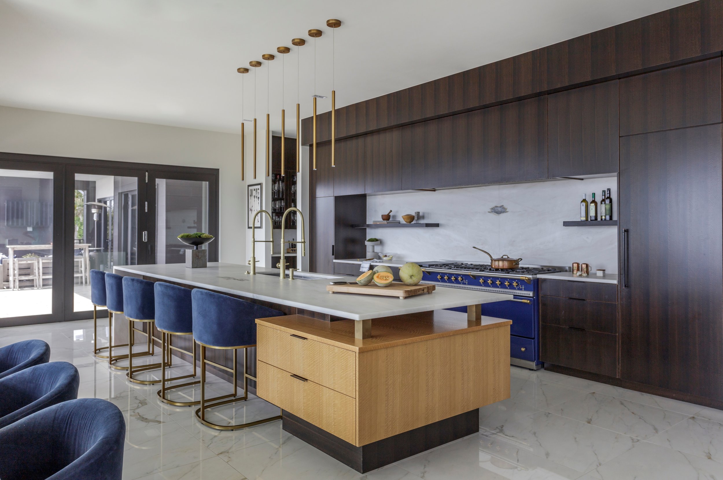 Modern kitchen with dark wood cabinets, a white marble backsplash, and a large island with a white countertop and blue velvet chairs. There are hanging gold pendant lights above the island. The kitchen has glass sliding doors leading outside, and a s