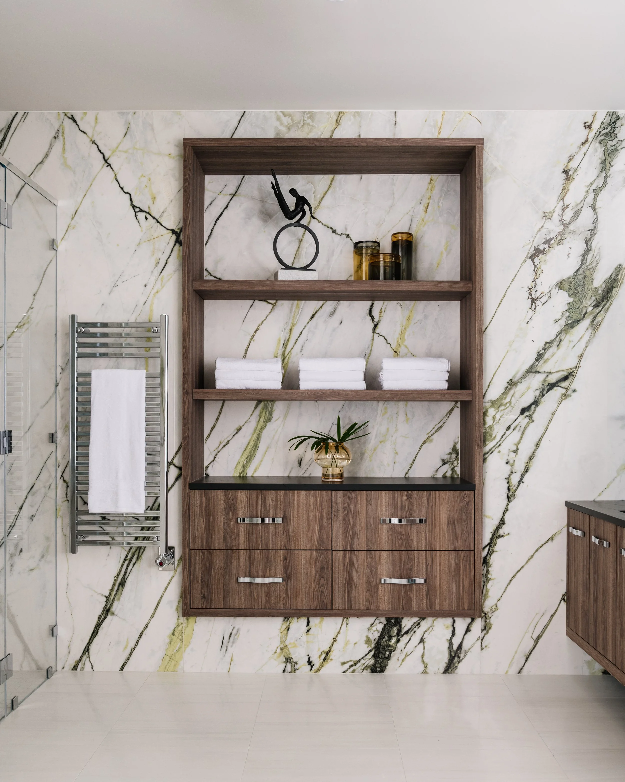 Modern bathroom with a marble wall behind a wooden cabinet, a decorative sculpture of a gymnast on a ring, three amber-colored candles, neatly folded white towels, a plant in a glass vase, and a chrome towel radiator to the left.