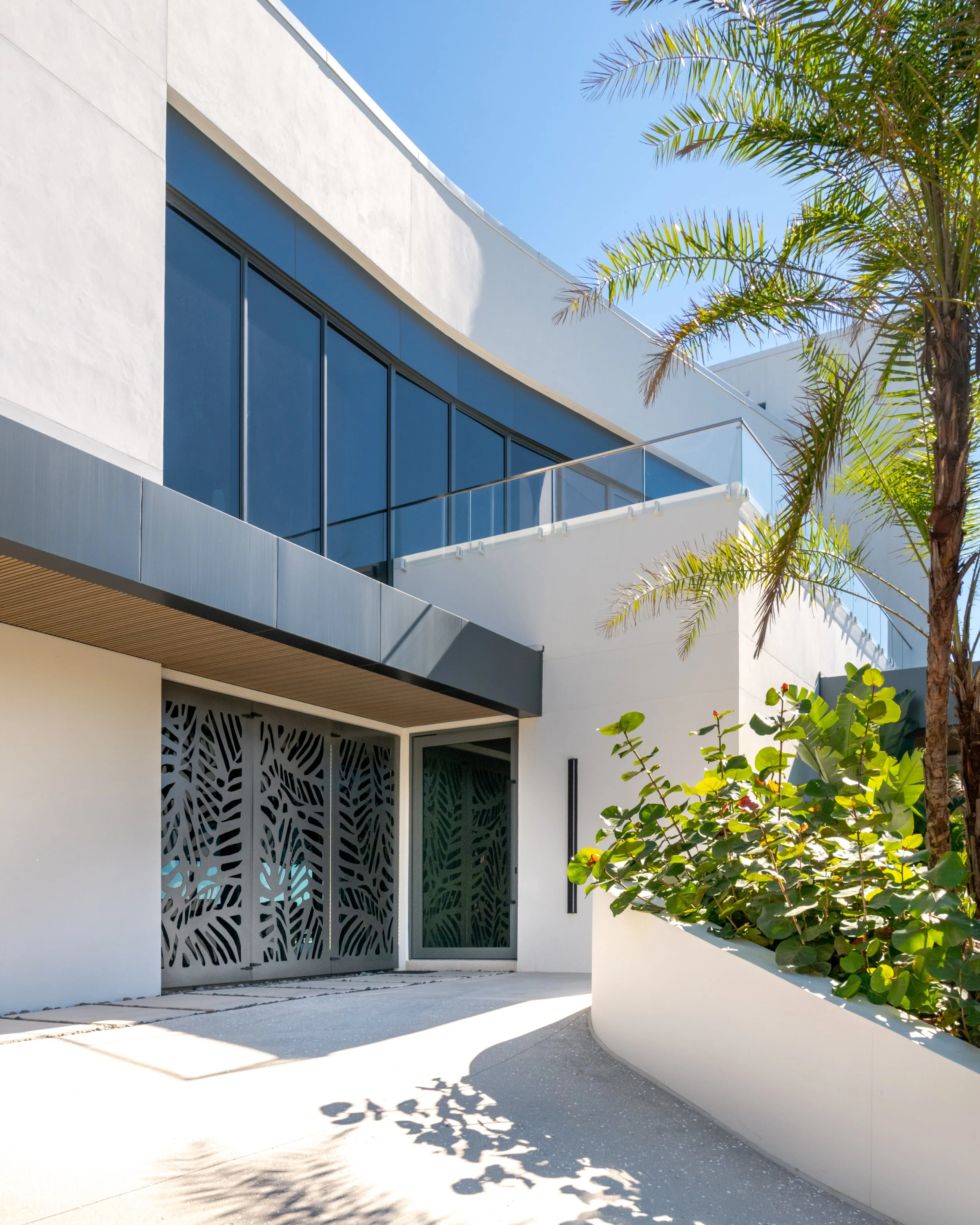 Modern residential building with white exterior walls, large glass windows, and a balcony with a glass railing. There are green palm trees and plants nearby, and the sky is clear and blue.
