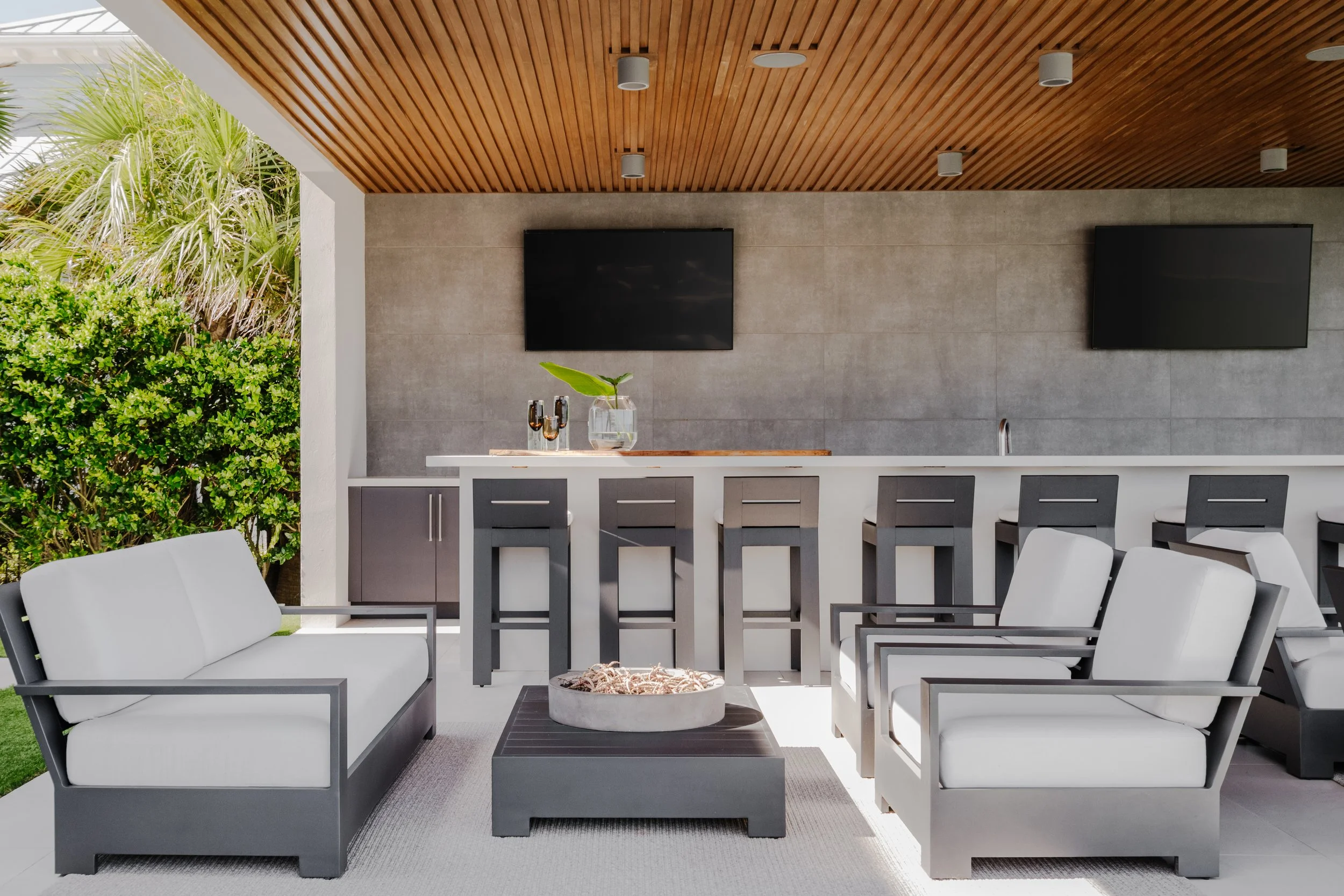 Outdoor patio seating area with white cushioned chairs, a high-top table, a potted plant, and a wood-paneled ceiling. Two large flat-screen TVs mounted on the wall and greenery visible on the left side.