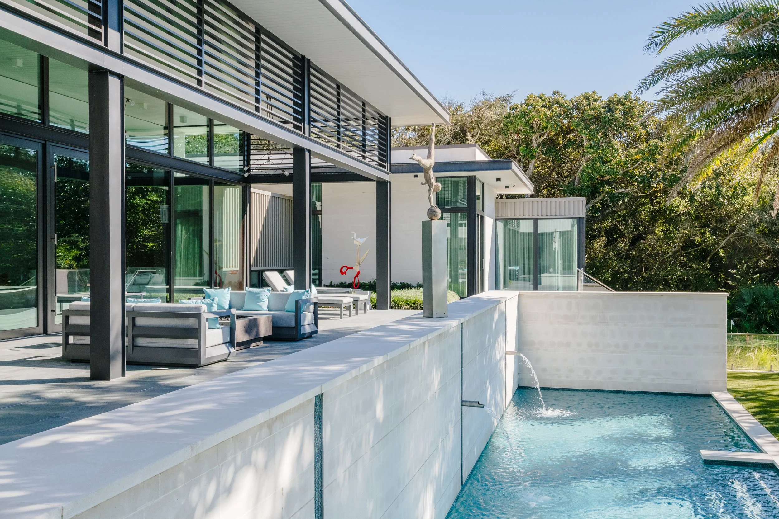 Modern house with large glass windows and a terrace, next to a swimming pool with water spouts, surrounded by lush green trees.