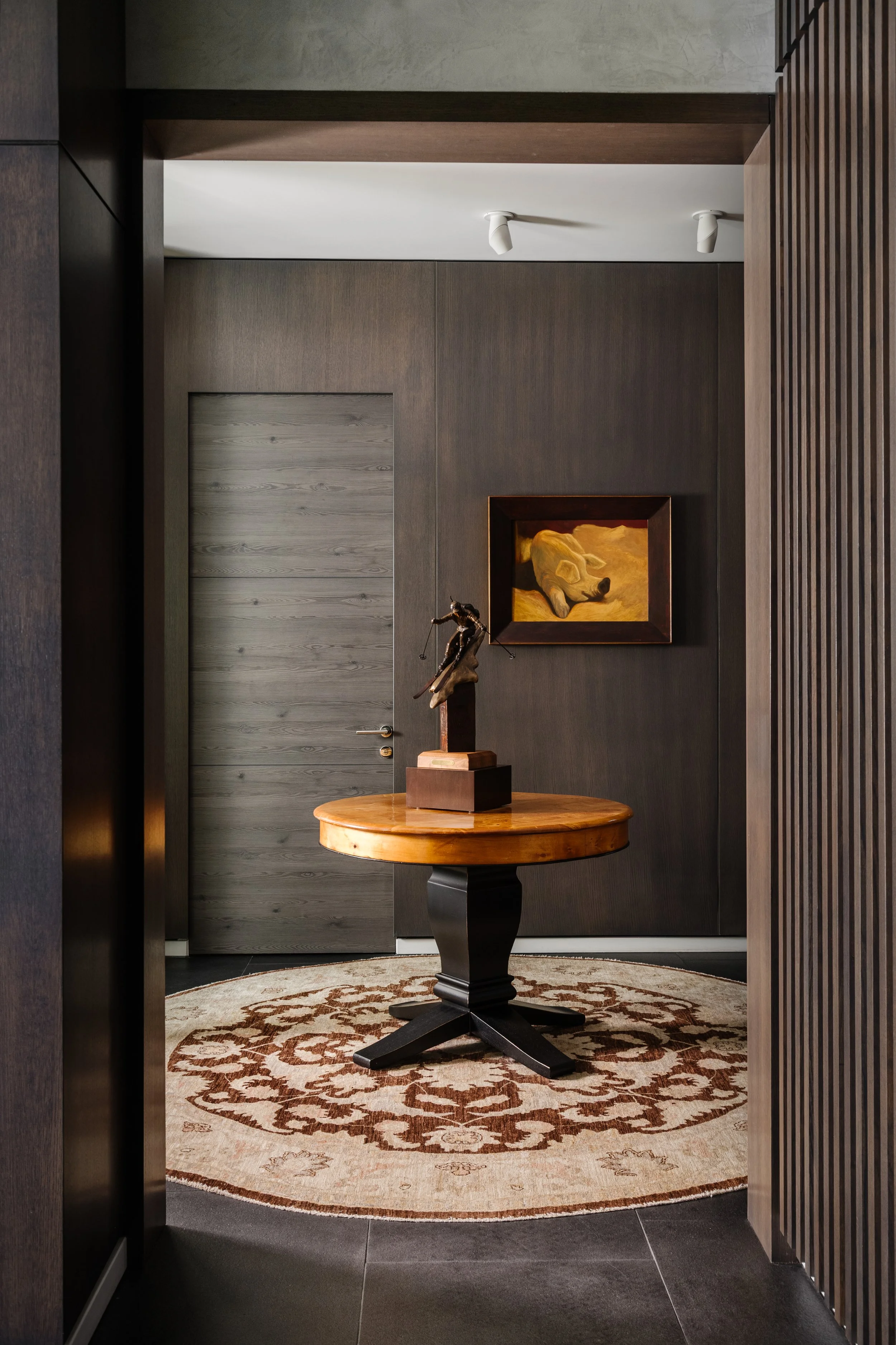 Interior view of a modern room with dark walls, a round wooden table with a sculpture on top, a patterned rug, a door, and a framed painting of a dog on the wall.