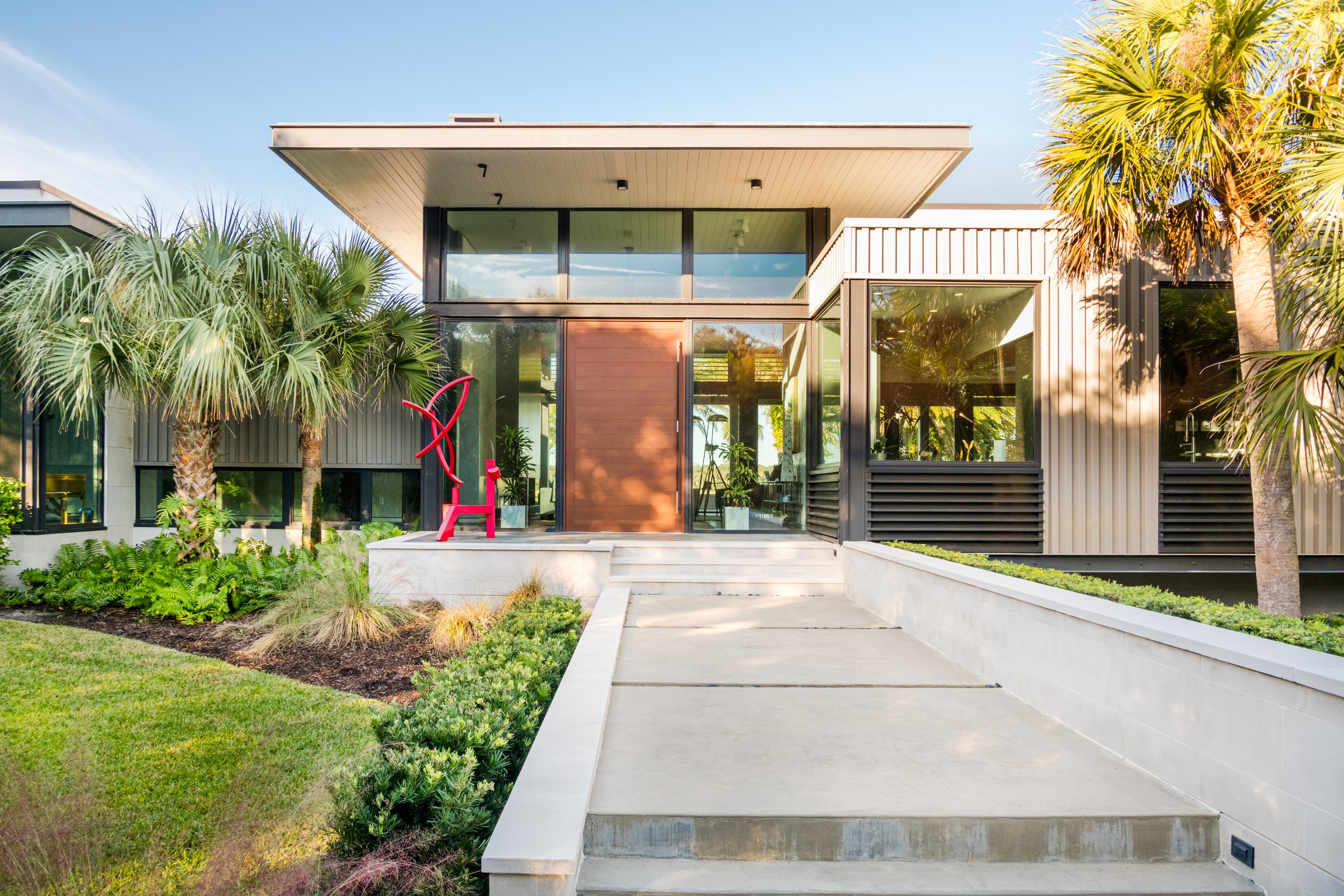 Modern two-story house with large glass windows, surrounded by lush green plants and trees, with a concrete pathway leading to the front door.