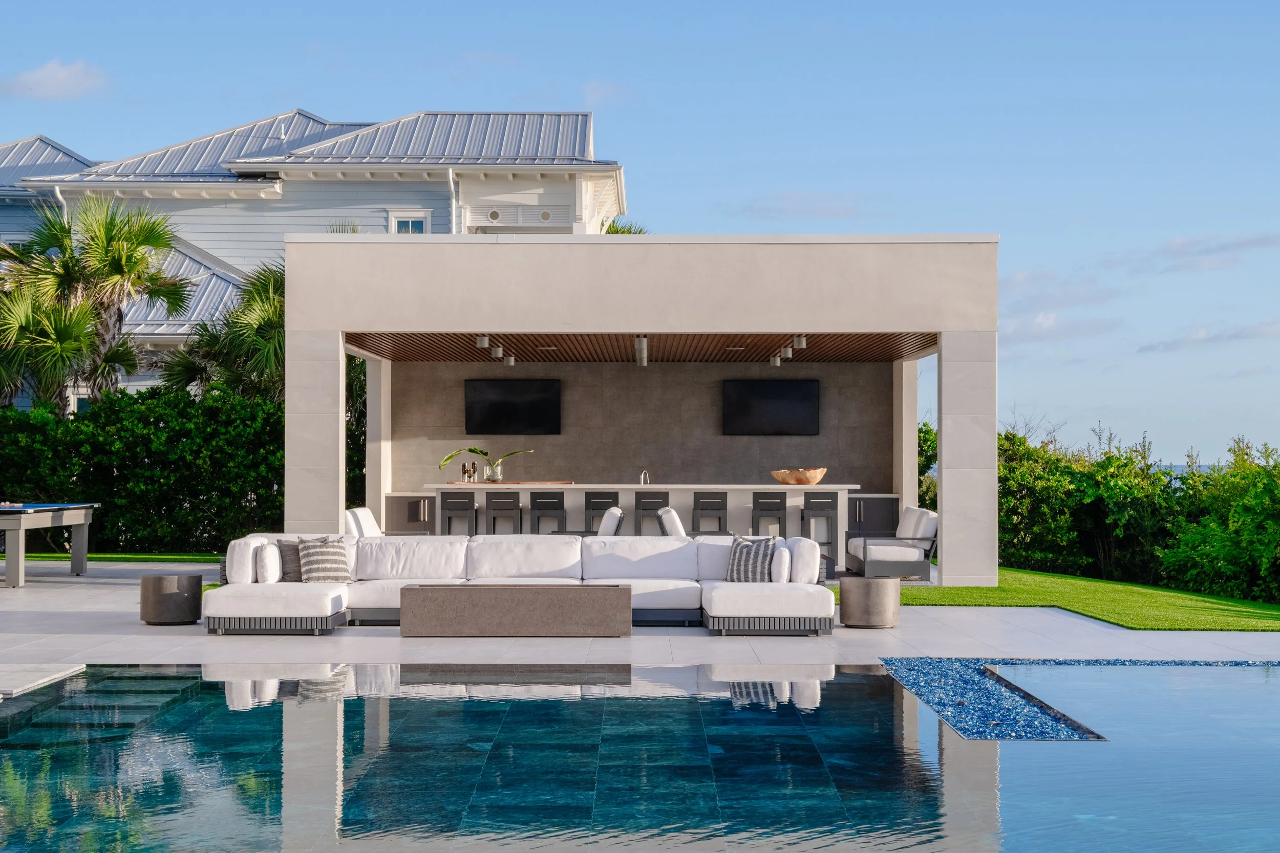 Luxury outdoor patio with pool, white sofa, bar area, and modern house in the background, under a clear blue sky.