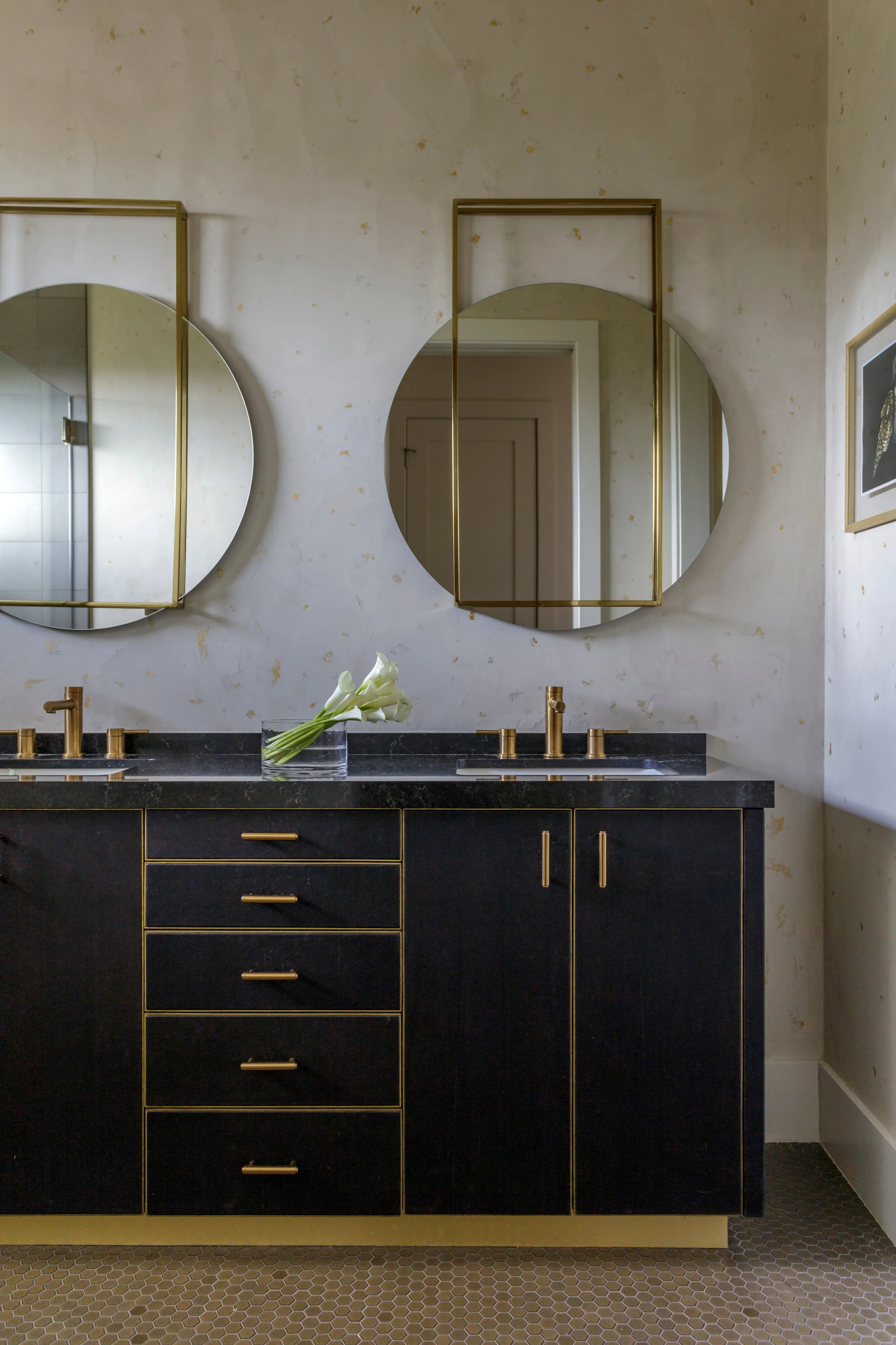 A bathroom vanity with a black finish, gold handles, and a gold trim, featuring three drawers and two cabinets. On top of the vanity, there is a clear glass vase with white calla lilies. Two round mirrors with gold frames are mounted on the wall abov