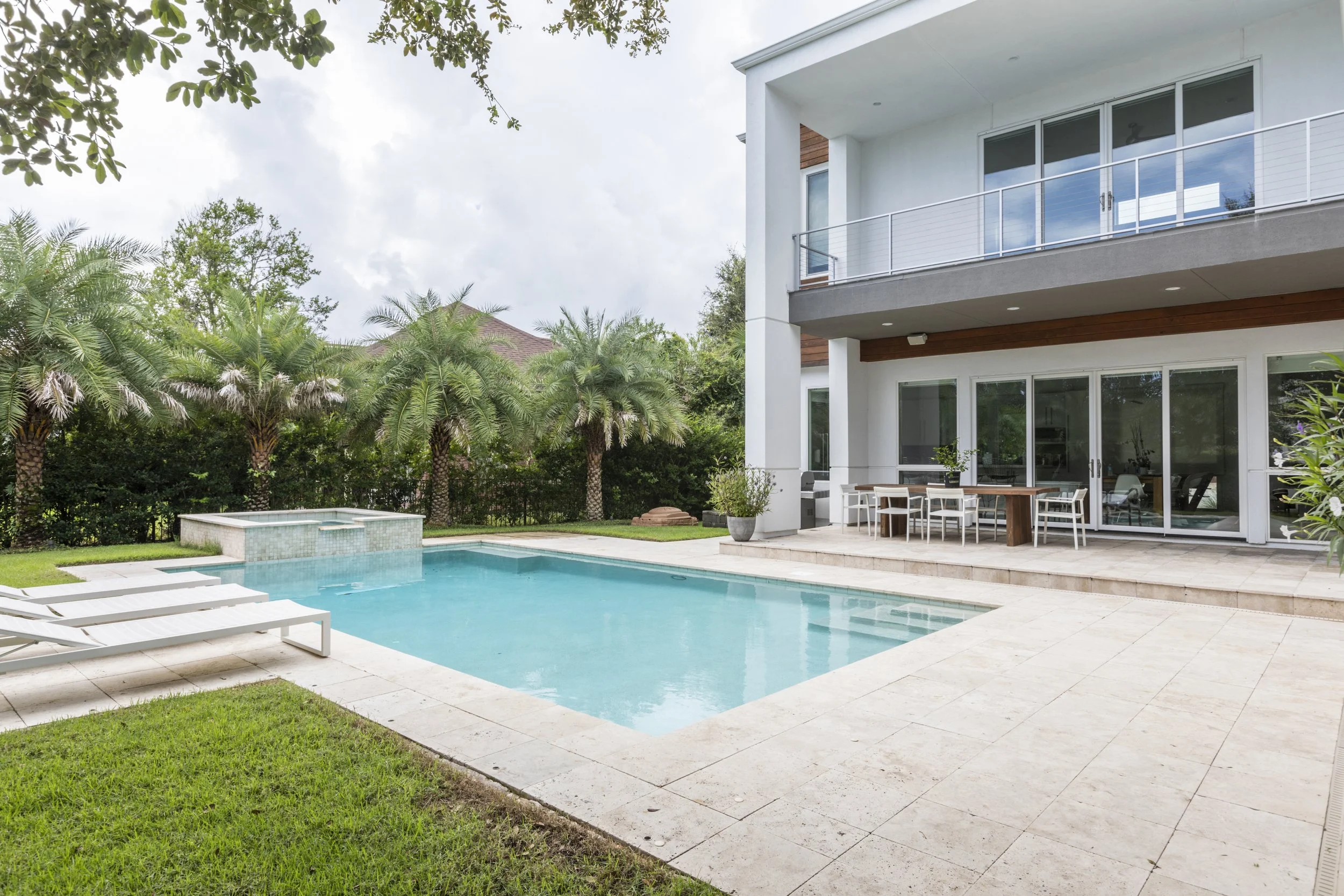 Modern house with a large swimming pool, outdoor dining area, surrounded by tall palm trees and lush greenery under an overcast sky.