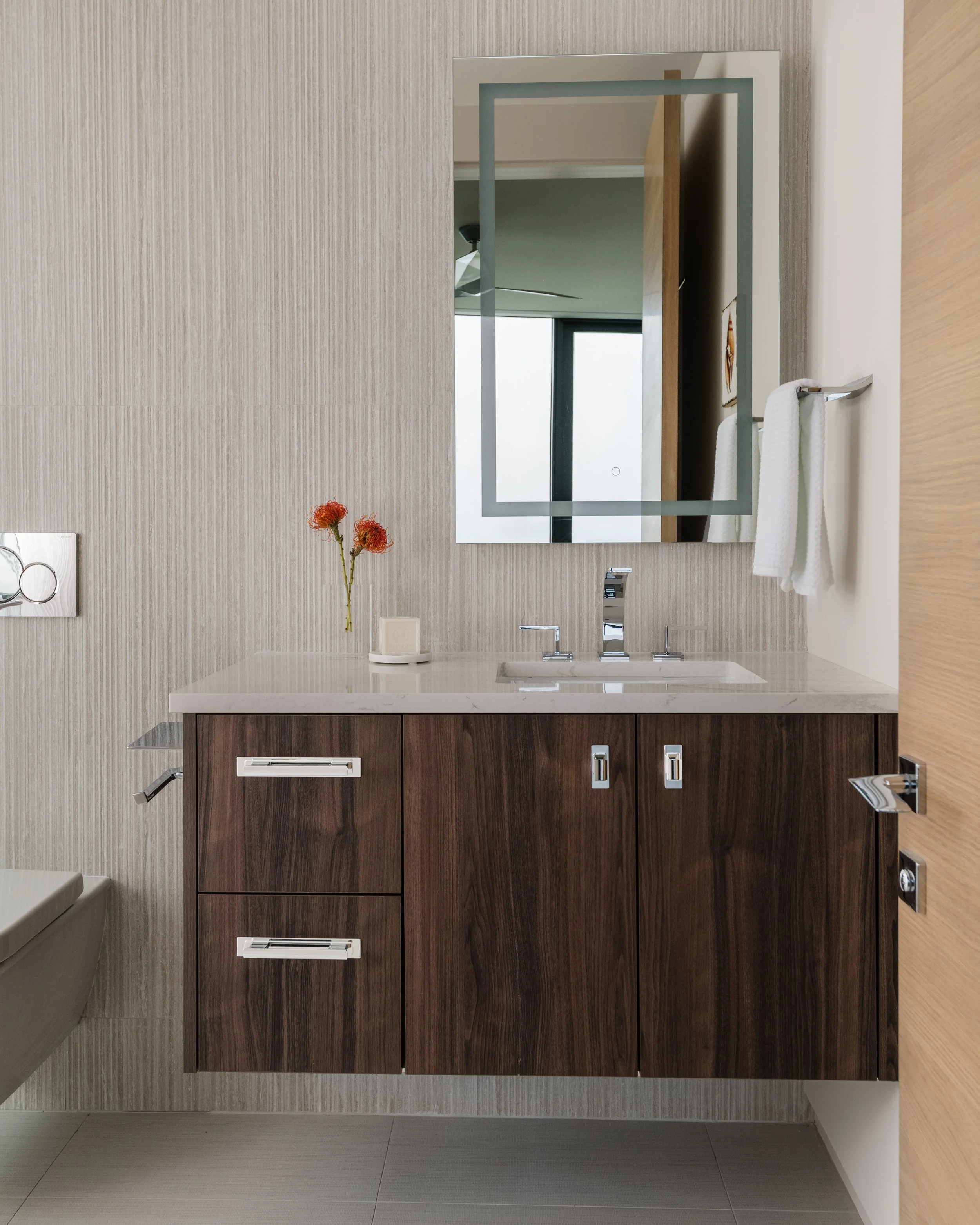 Modern bathroom with beige textured wall, large mirror, white towels on a metal towel rack, a wooden vanity with a marble countertop, a small white cylindrical decorative object, and a vase with orange flowers.