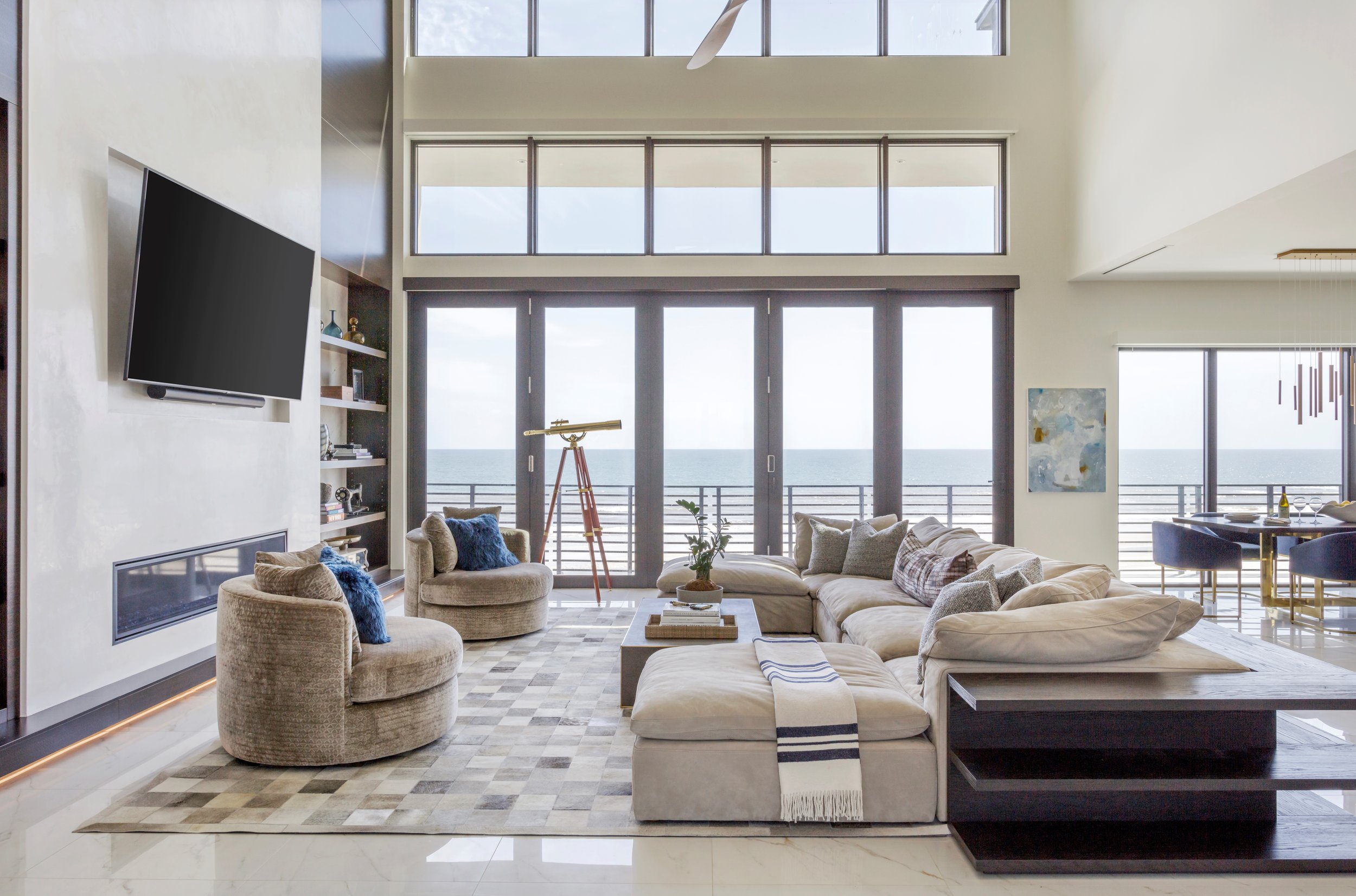 Modern living room with large windows overlooking the ocean, beige sectional sofa, armchairs, a patterned rug, and decorative items.