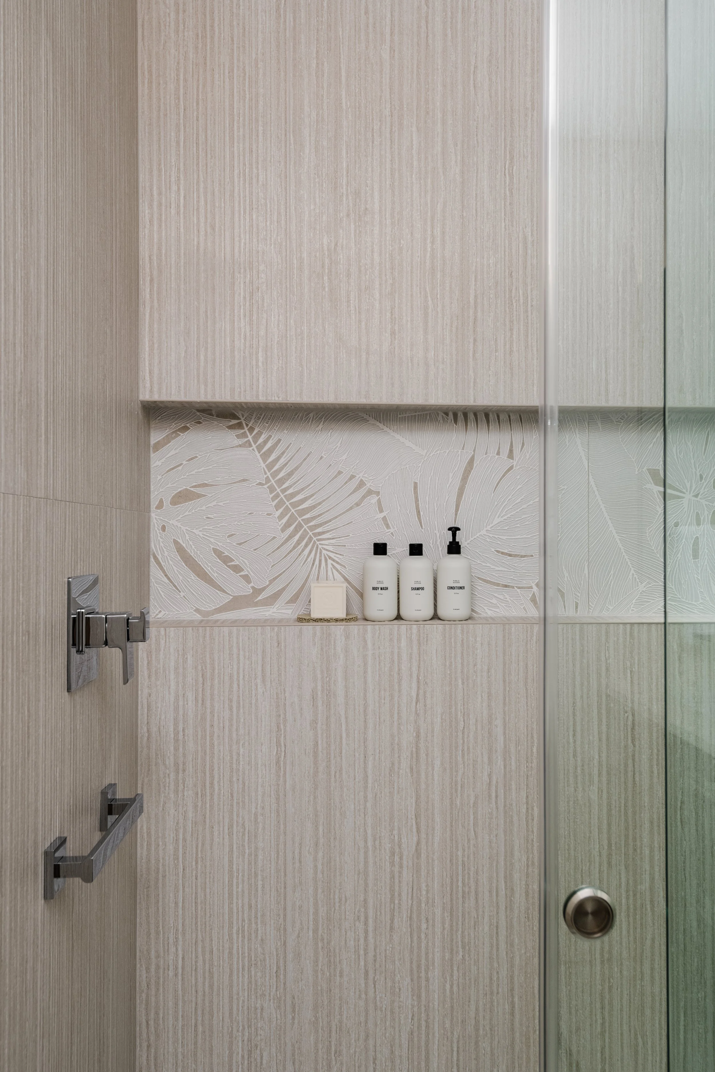 A bathroom shower door with a beige wood-textured wall. Inside the shower, there is a built-in shelf holding three white bottles labeled body wash, shampoo, and conditioner, along with a small soap dish.