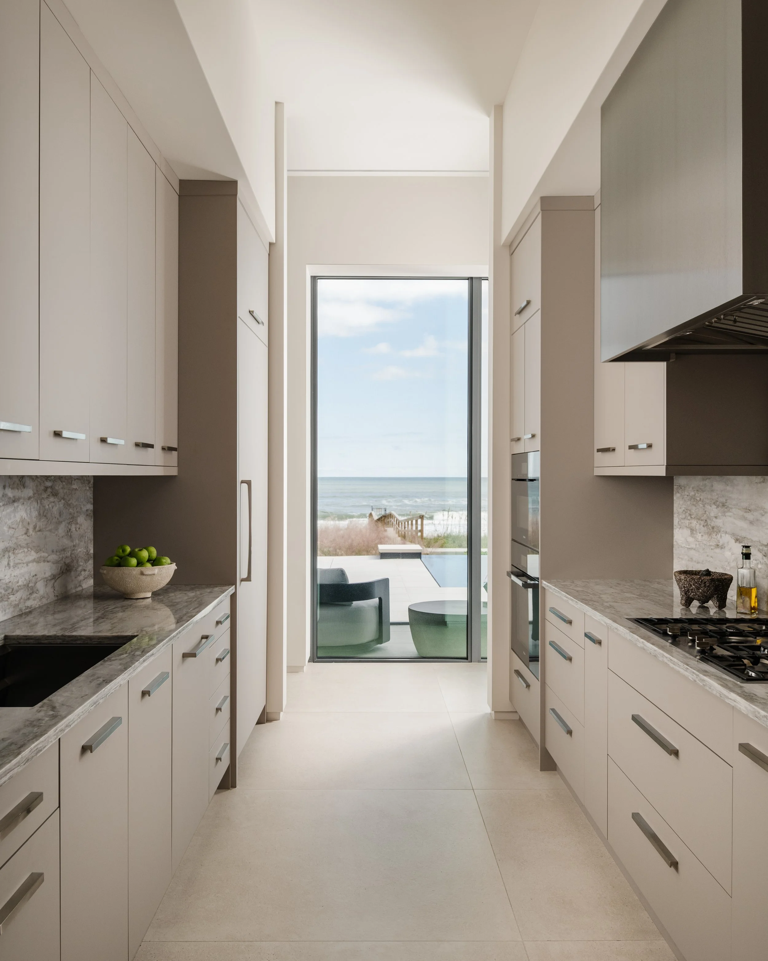 Modern kitchen with white cabinets, granite countertops, stainless steel appliances, and a view of the beach and ocean through a large glass door.