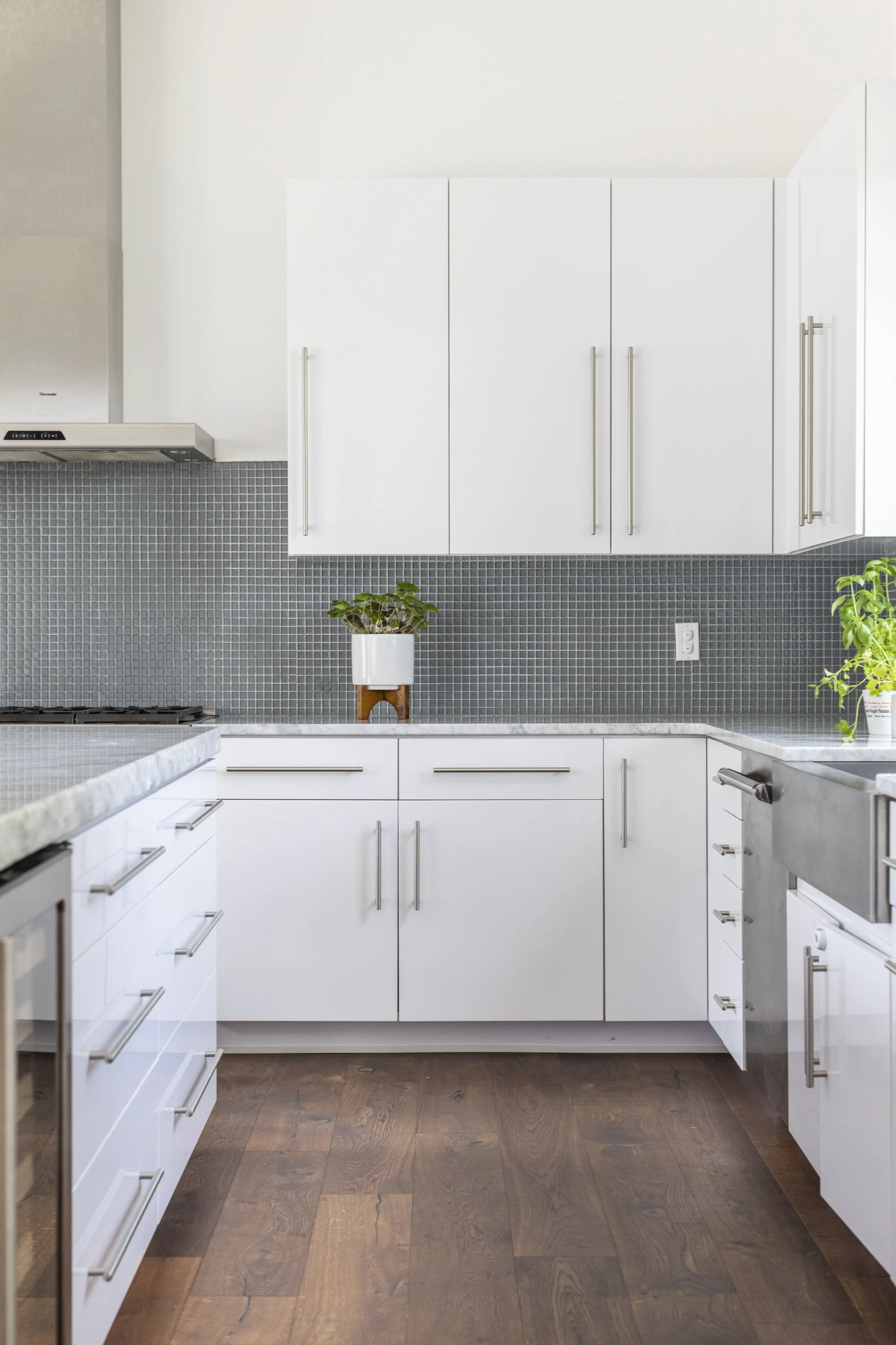 White kitchen cabinets with silver handles, gray mosaic tile backsplash, dark hardwood floor, and a small potted plant on a wooden stand.