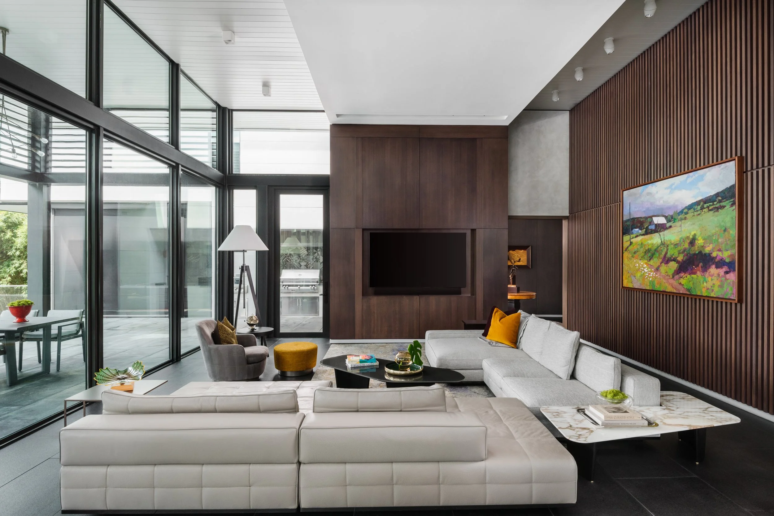 Modern living room with large glass windows, white sectional sofa, colorful throw pillows, black coffee table with books, yellow ottomans, and a wall mounted TV. Artwork on a wooden slatted wall, indoor plants, and a glass dining area in the backgrou