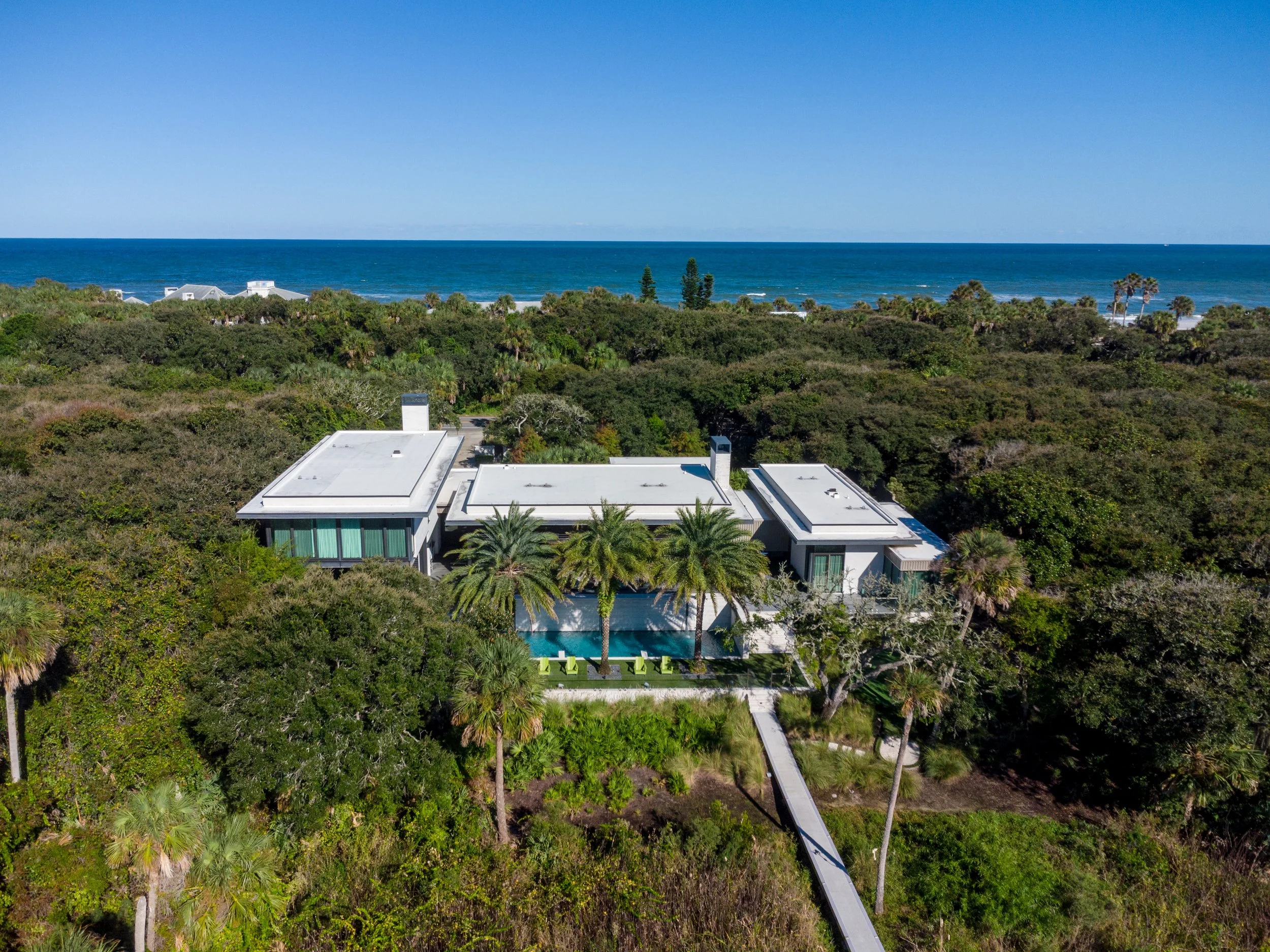 Aerial view of a beachfront house surrounded by lush greenery and palm trees with the ocean in the background under a clear blue sky.