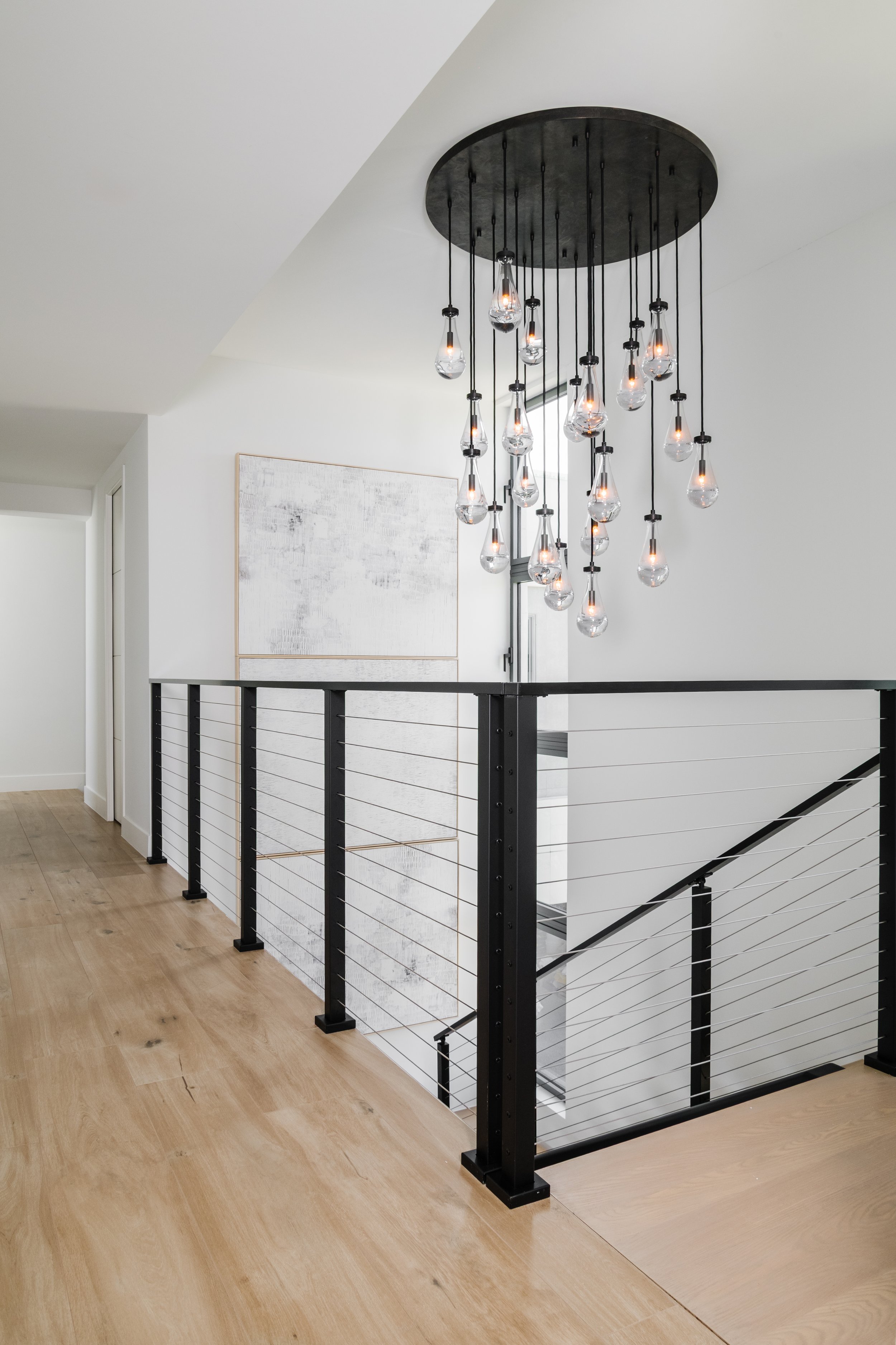 Modern chandelier with hanging glass bulbs in a minimalist interior space with white walls and wooden flooring.