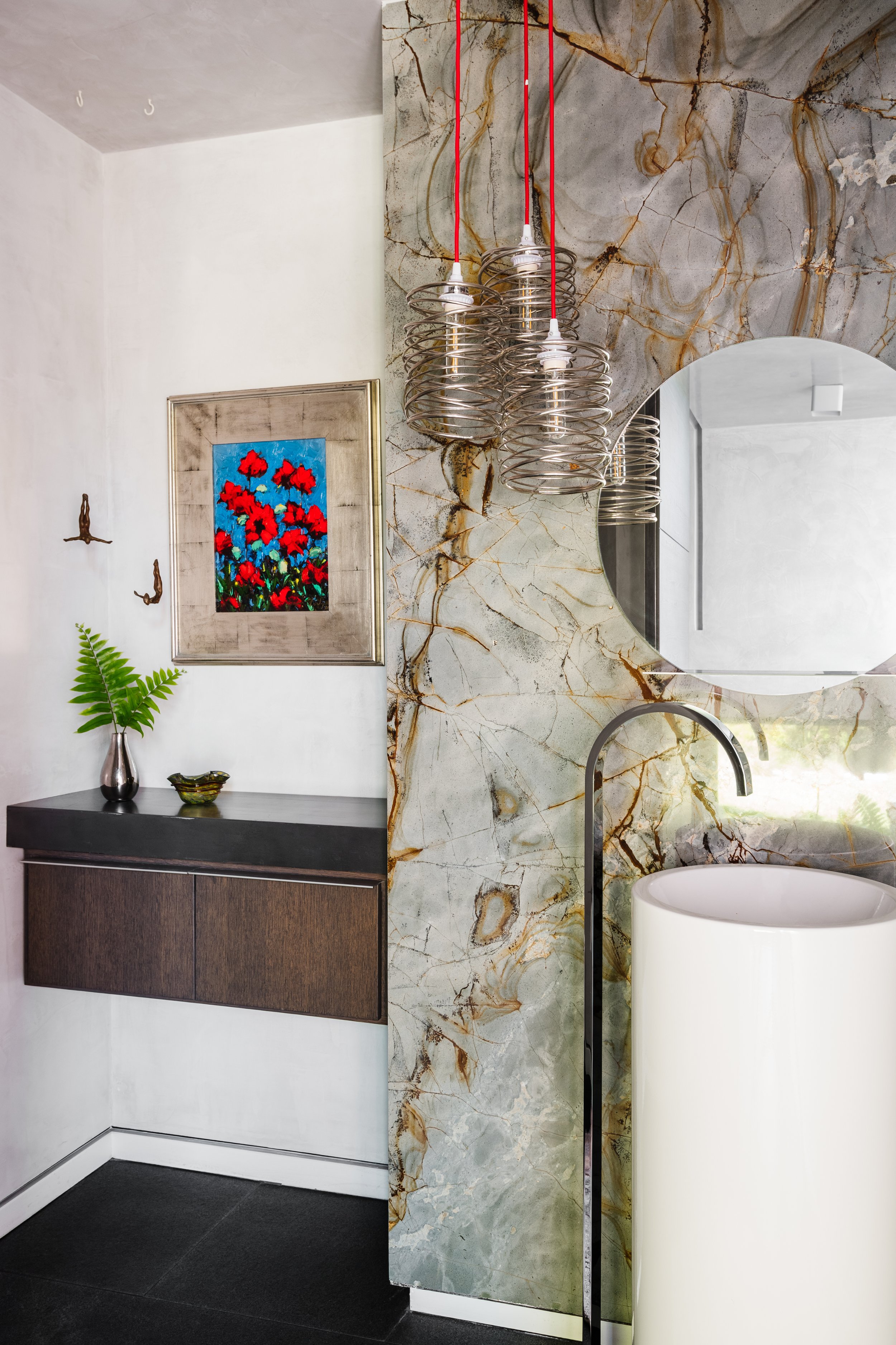 Interior view of a modern bathroom with a marble wall, round mirror, and a white vessel sink. A decorative framed artwork of red flowers with a blue background hangs on a white wall, with a vase and small bowl below it. Hanging from the ceiling are t