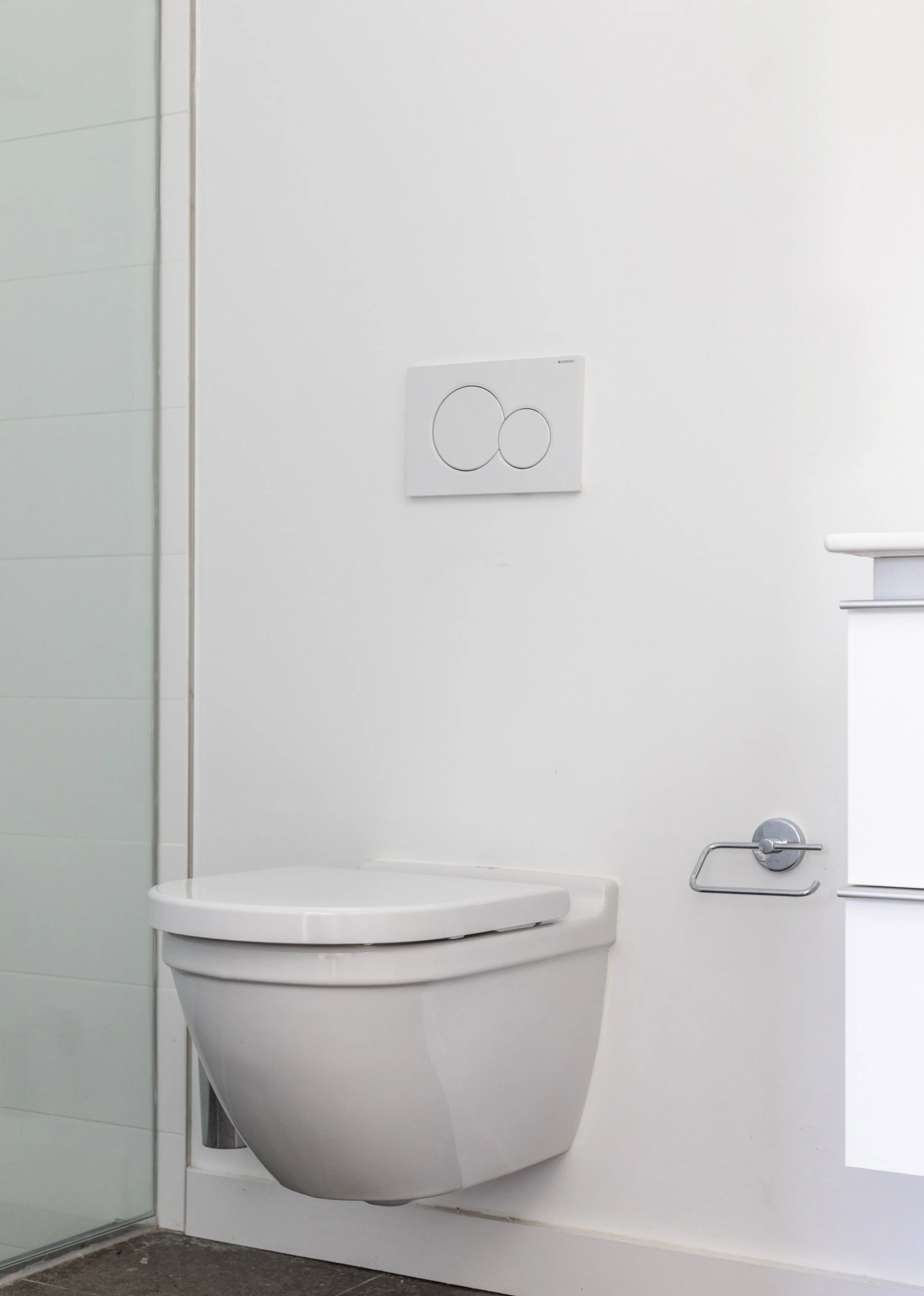 Public restroom with a wall-mounted toilet, flush plate, and toilet paper holder on the wall. Part of a walk-in shower with glass door is visible on the left. The floor is tiled with dark tiles.