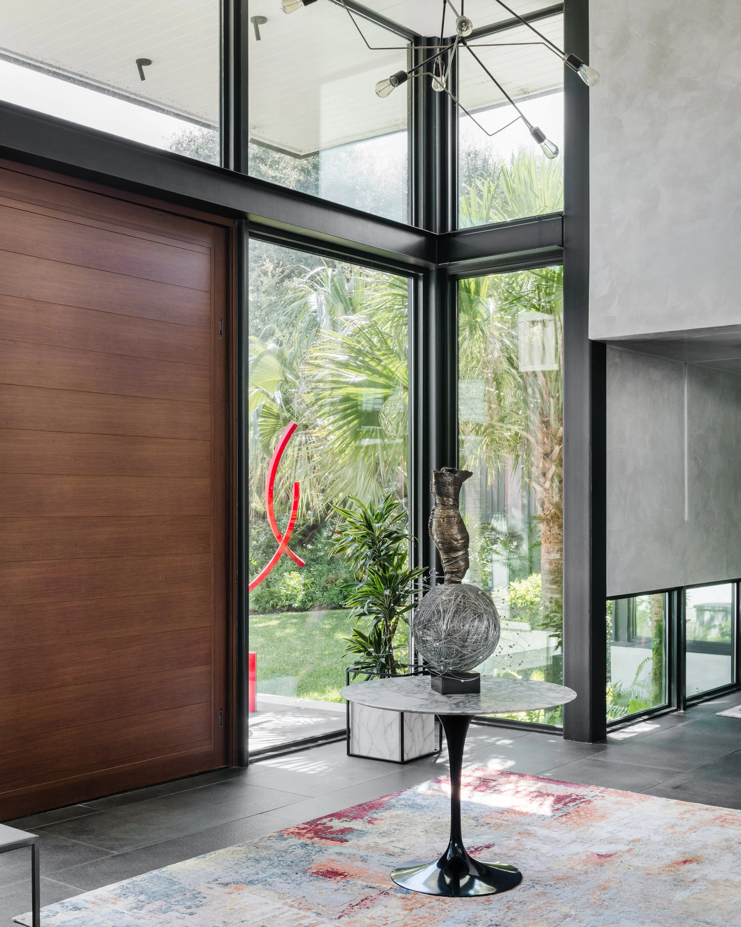 Modern interior with glass walls showing outdoor greenery, a marble-topped table with a decorative sphere sculpture, a plant, and a colorful area rug.
