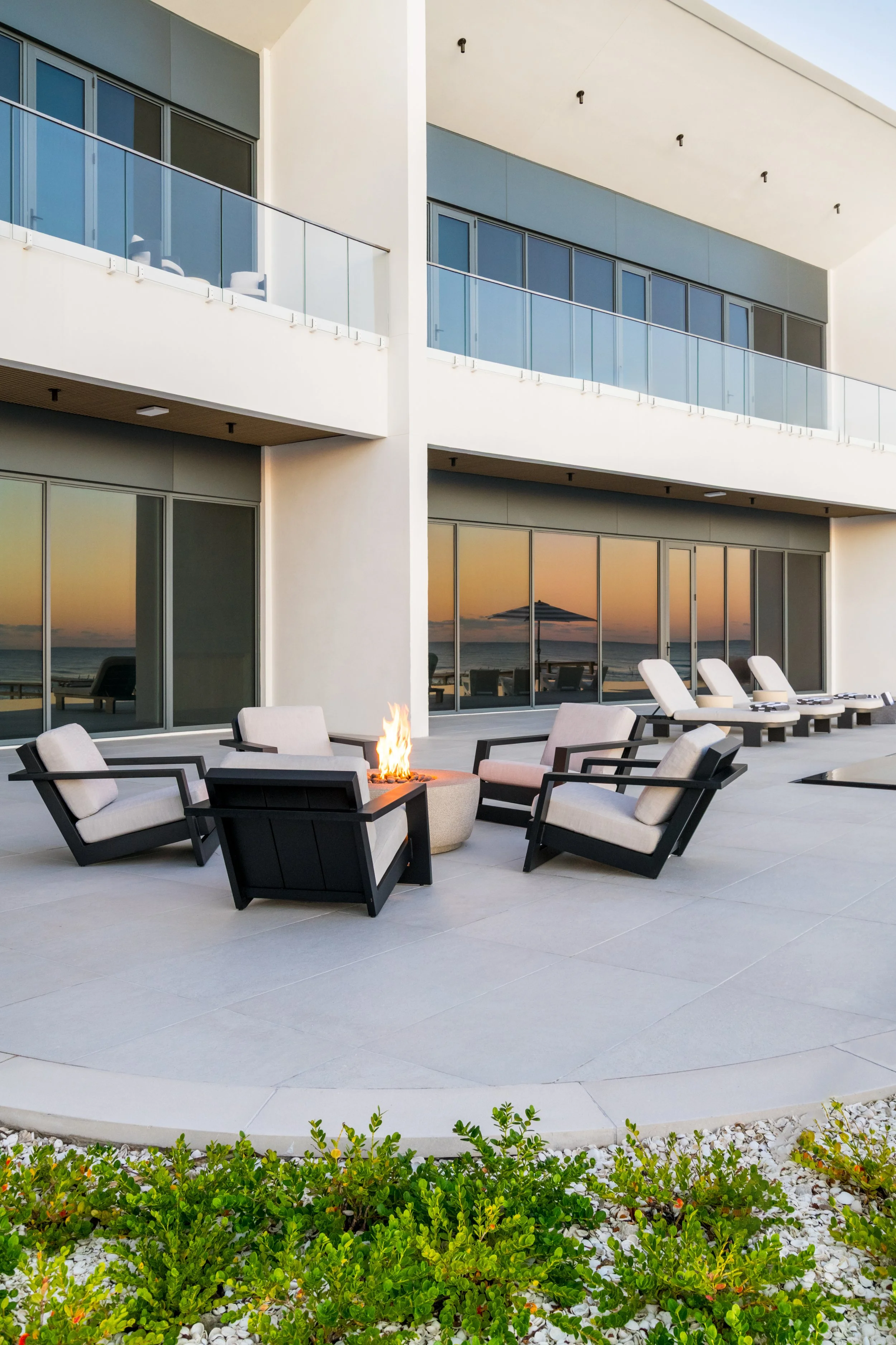 Modern outdoor patio area with lounge chairs, a fire pit, large glass windows reflecting sunset, and a view of the ocean with a beach umbrella in the background.