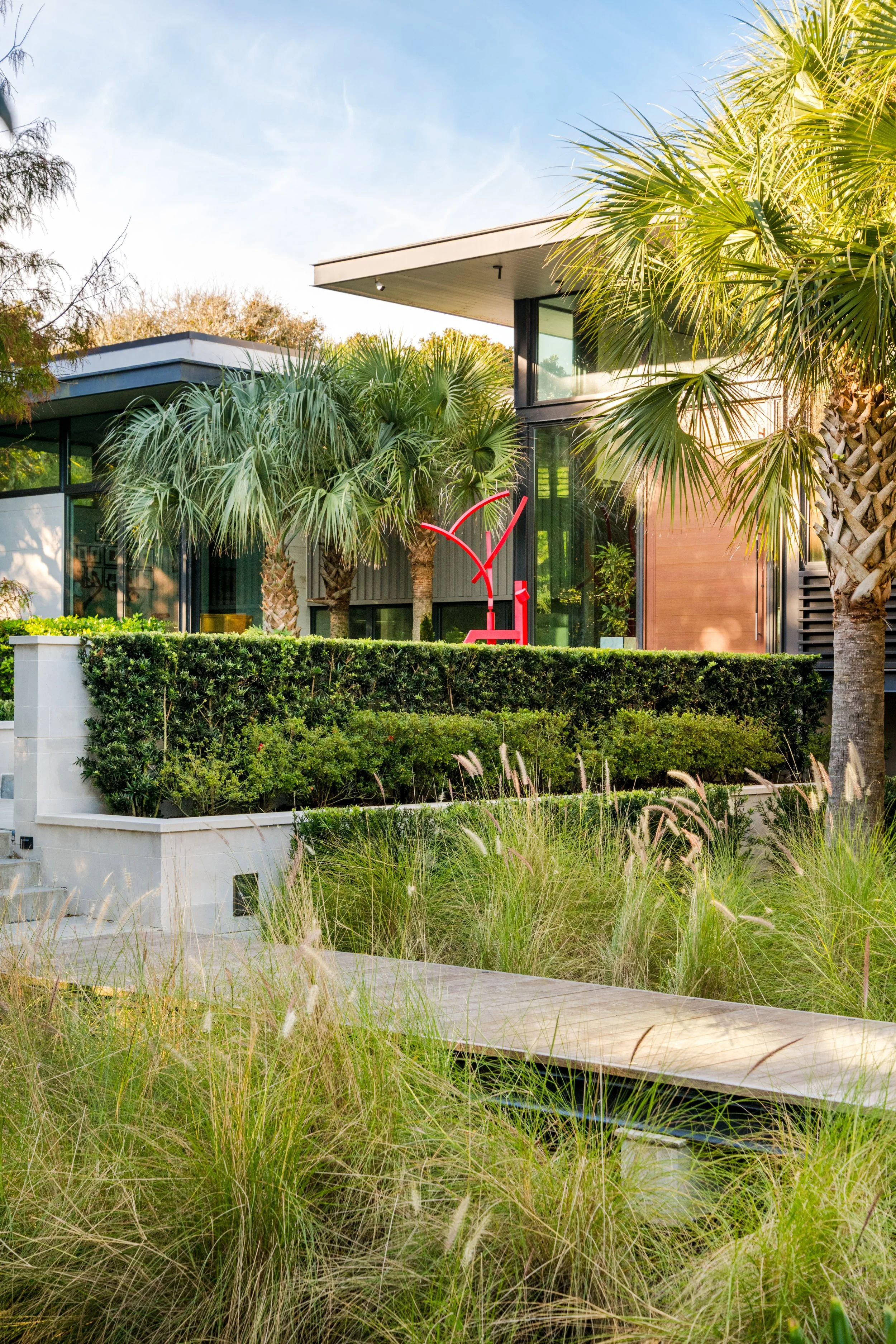 Modern house with large windows surrounded by tropical plants and palm trees, with a bright sky and some grass in the foreground.