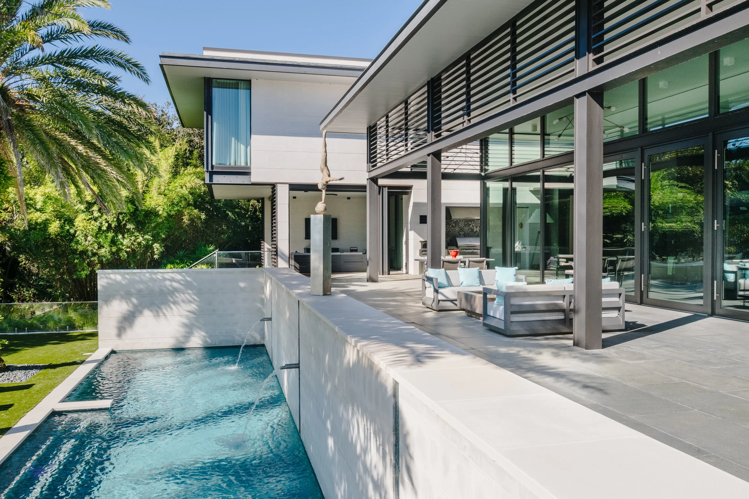 Modern house with a pool and outdoor seating area, surrounded by lush green trees and a palm tree, with a clear blue sky.