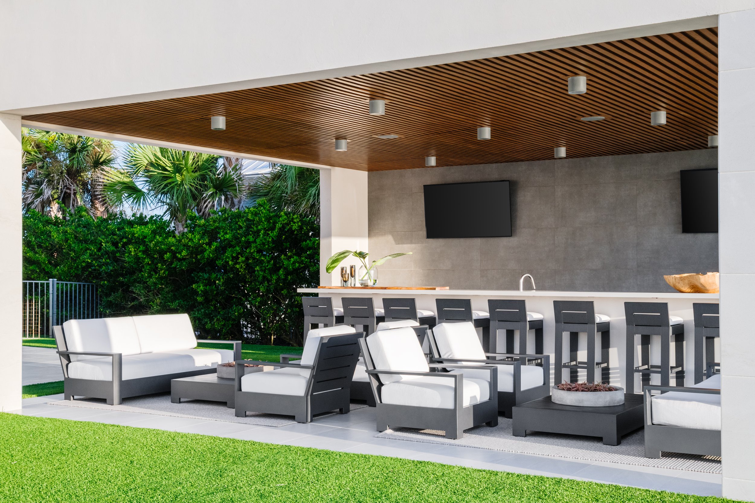 Modern outdoor patio with white and dark gray furniture, a bar with high chairs, and a wooden ceiling, surrounded by lush green plants.