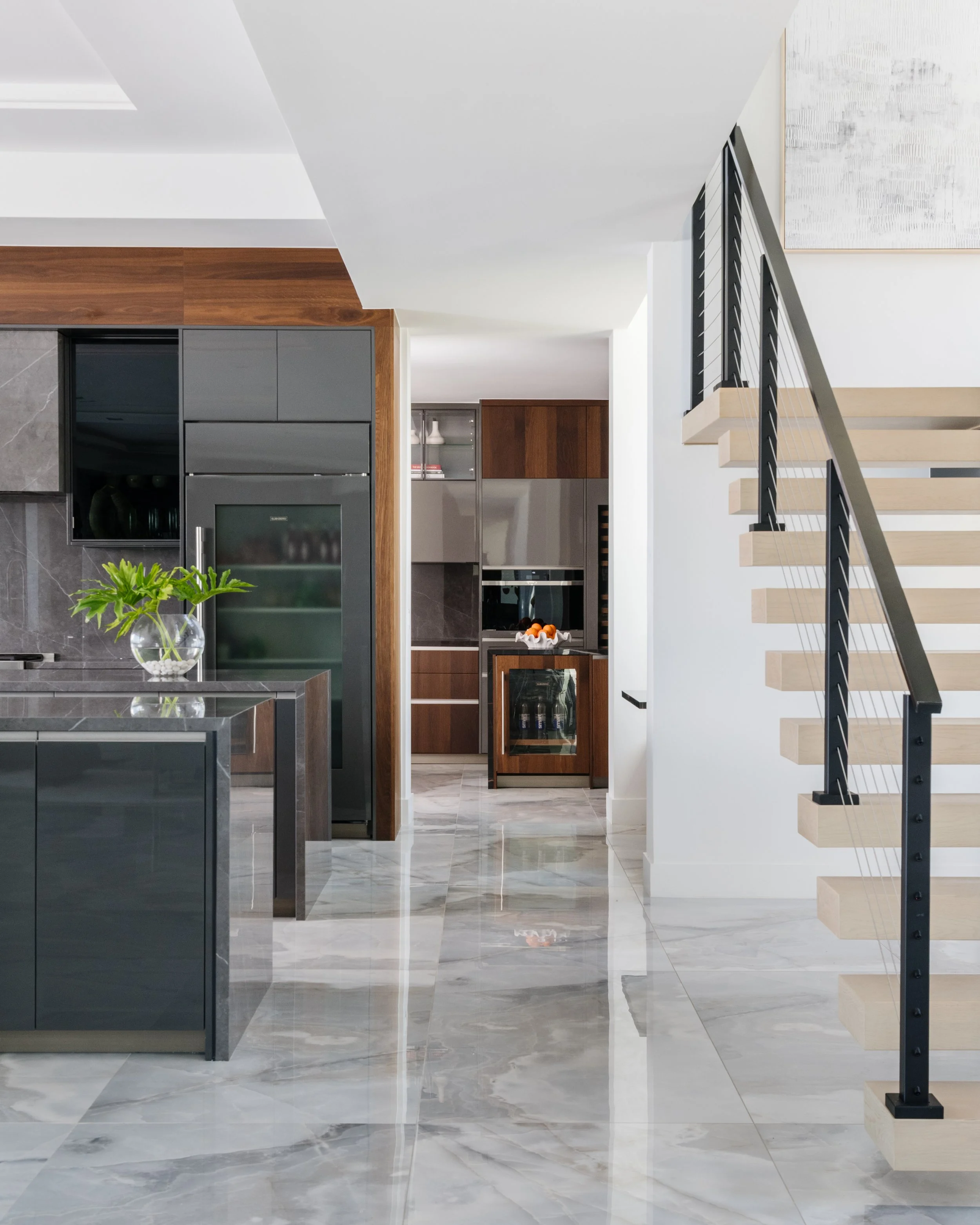 Modern interior of a house with black and wooden kitchen cabinets, a staircase with light wooden steps and black metal railing, white walls, and a marble floor.
