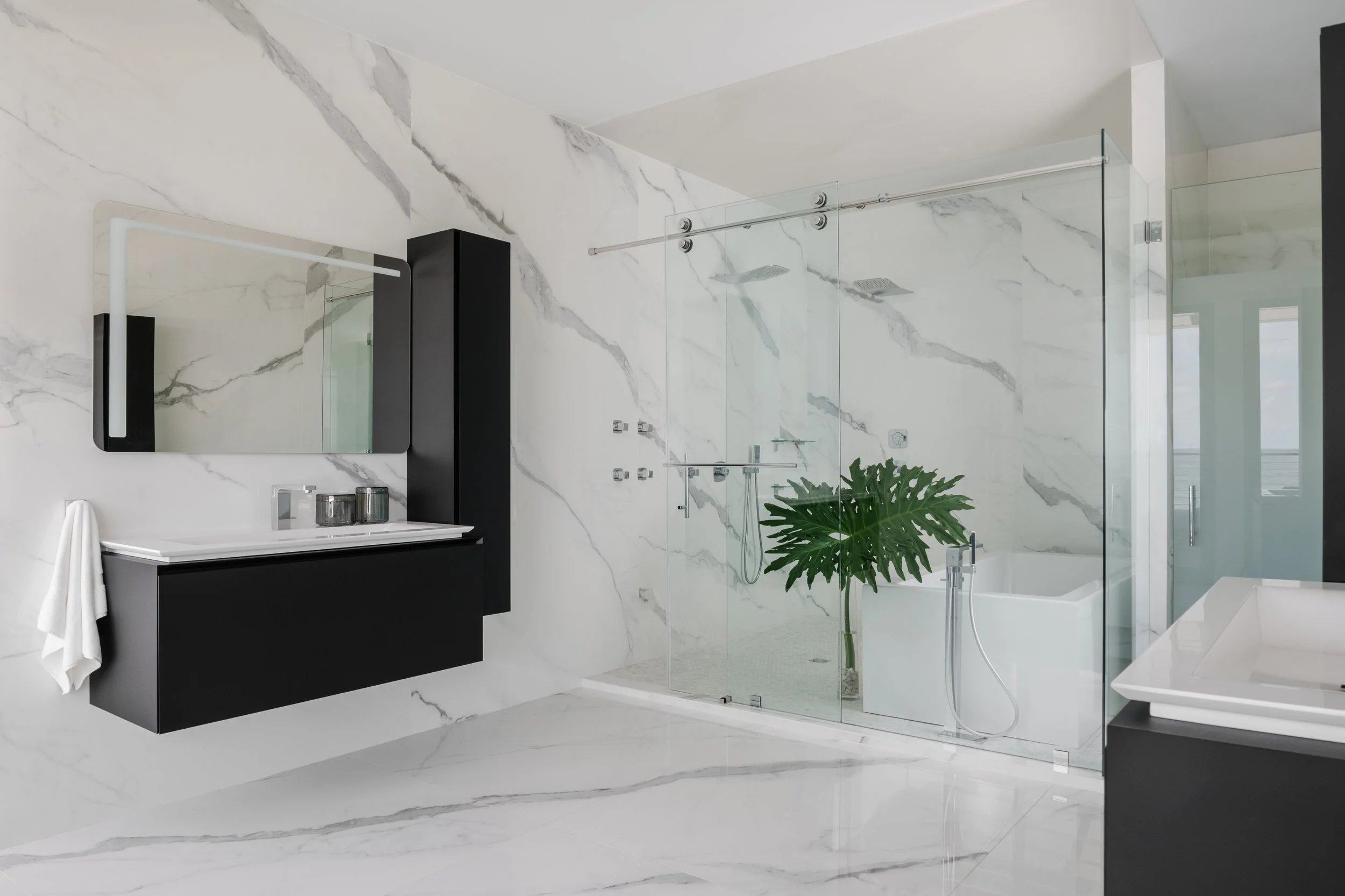 Modern bathroom with white marble walls and floors, black vanity with a mirror, glass shower enclosure with rainfall showerheads, white bathtub, and a green potted plant.