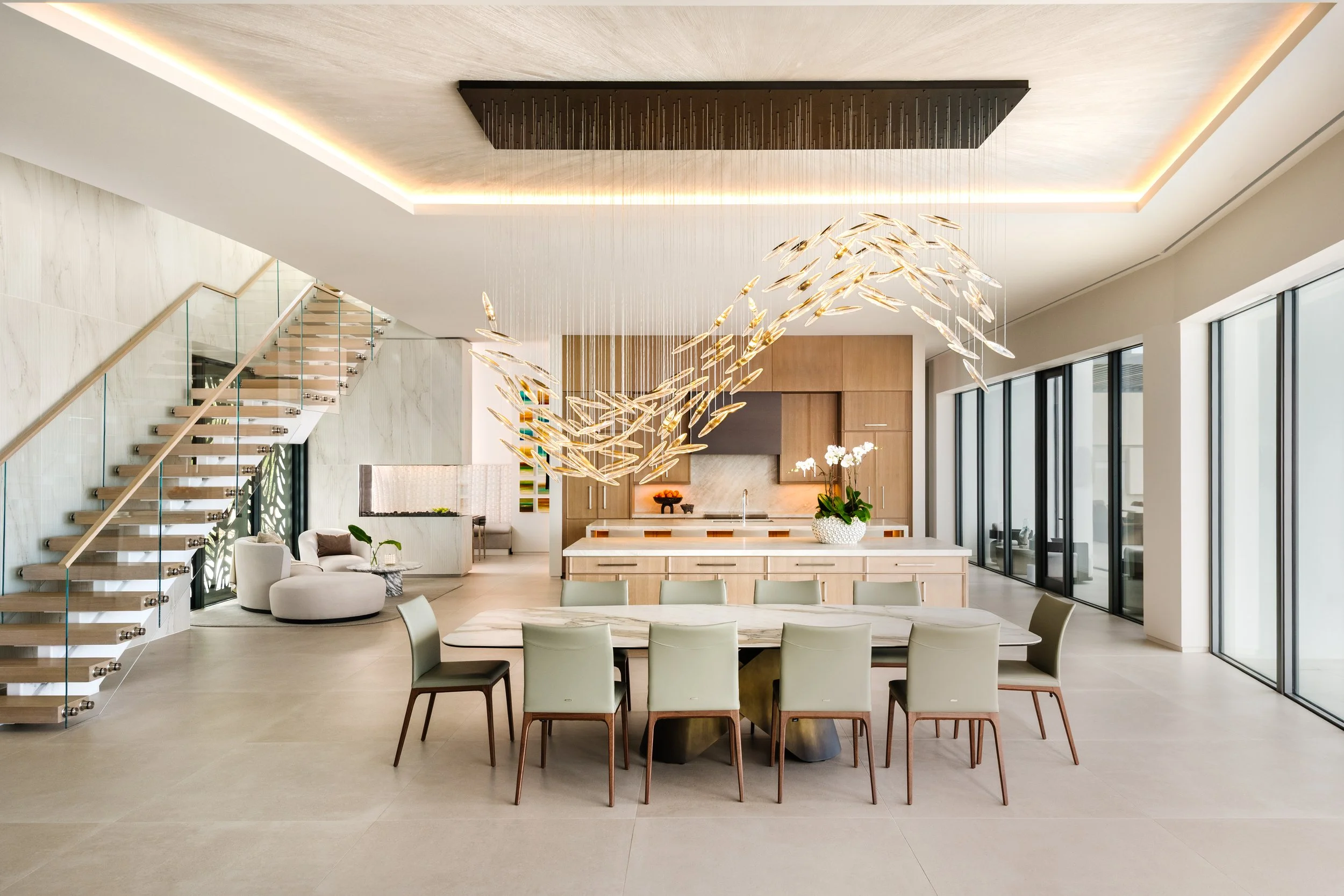 Open concept kitchen and dining area with modern decor, a chandelier resembling a school of fish, staircase with glass railing, large windows, and a seating area with armchair and side table.