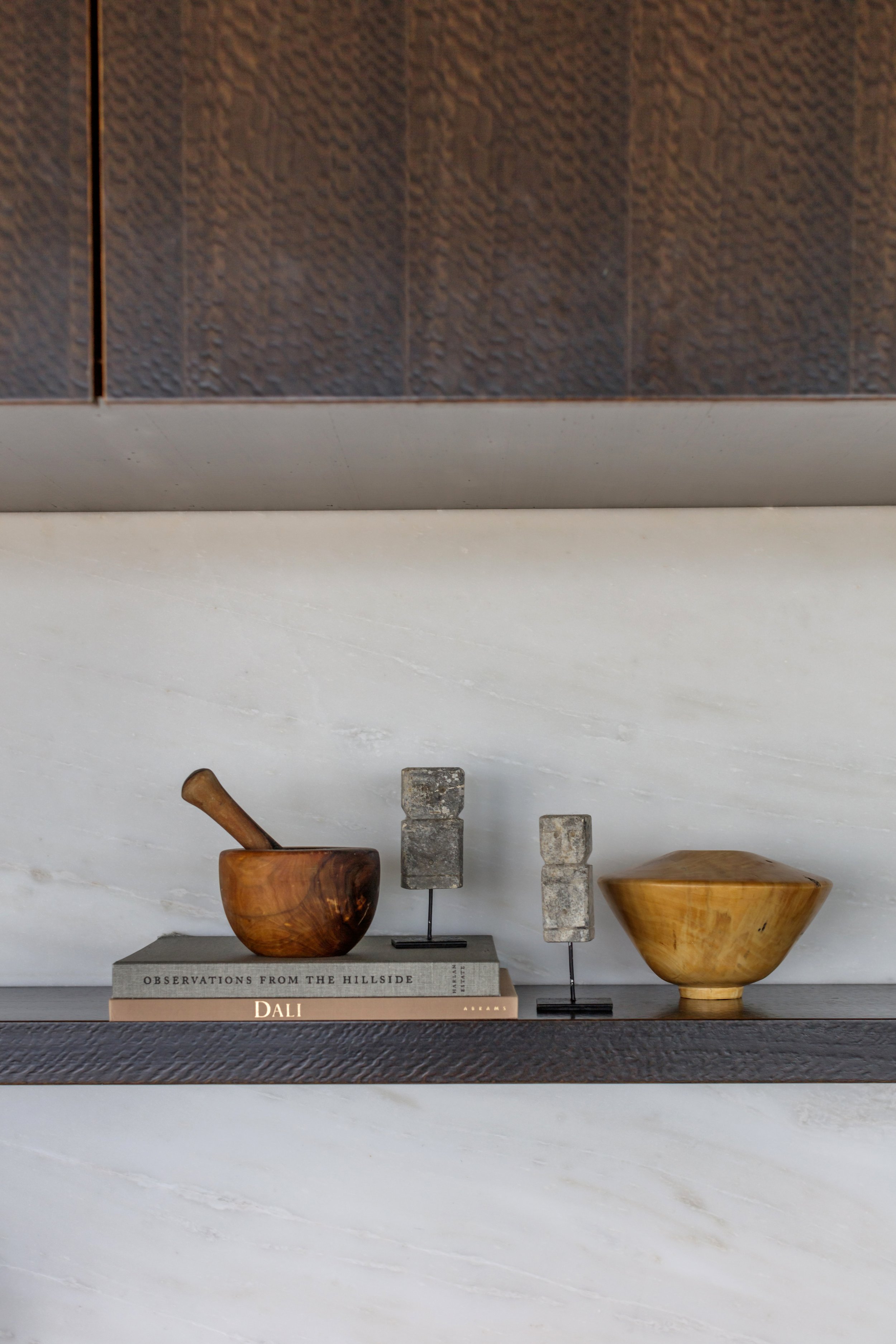 A bookshelf with three decorative bowls, a mortar and pestle, and books, including one titled "Observations from the Hillside" and another titled "DALI." The background features a partition with textured brown tiles.