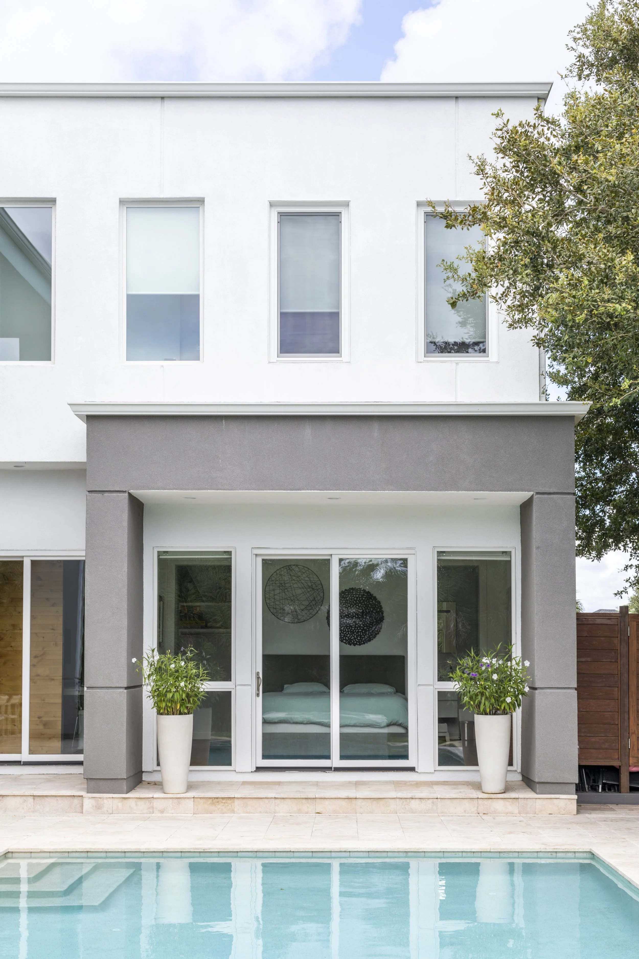 Modern two-story house with large glass doors opening to a pool, two potted plants on either side, and three tall windows on upper floor, surrounded by a tree and clear sky.