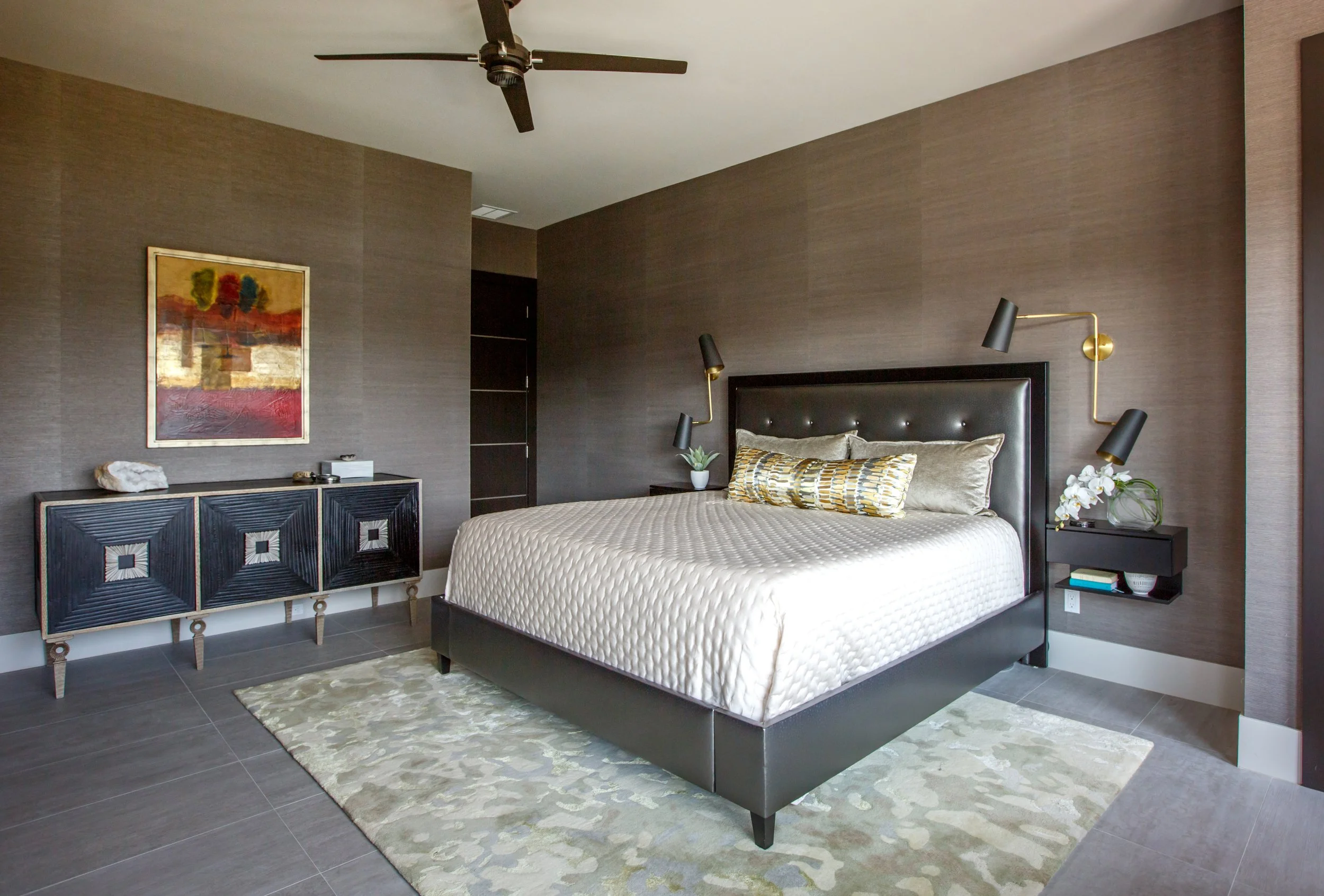 A modern bedroom with a queen-sized bed featuring a black tufted headboard, gold and black reading lamps mounted on either side, a small black bedside table with books, and a potted orchid. There's a colorful abstract painting on the taupe textured w