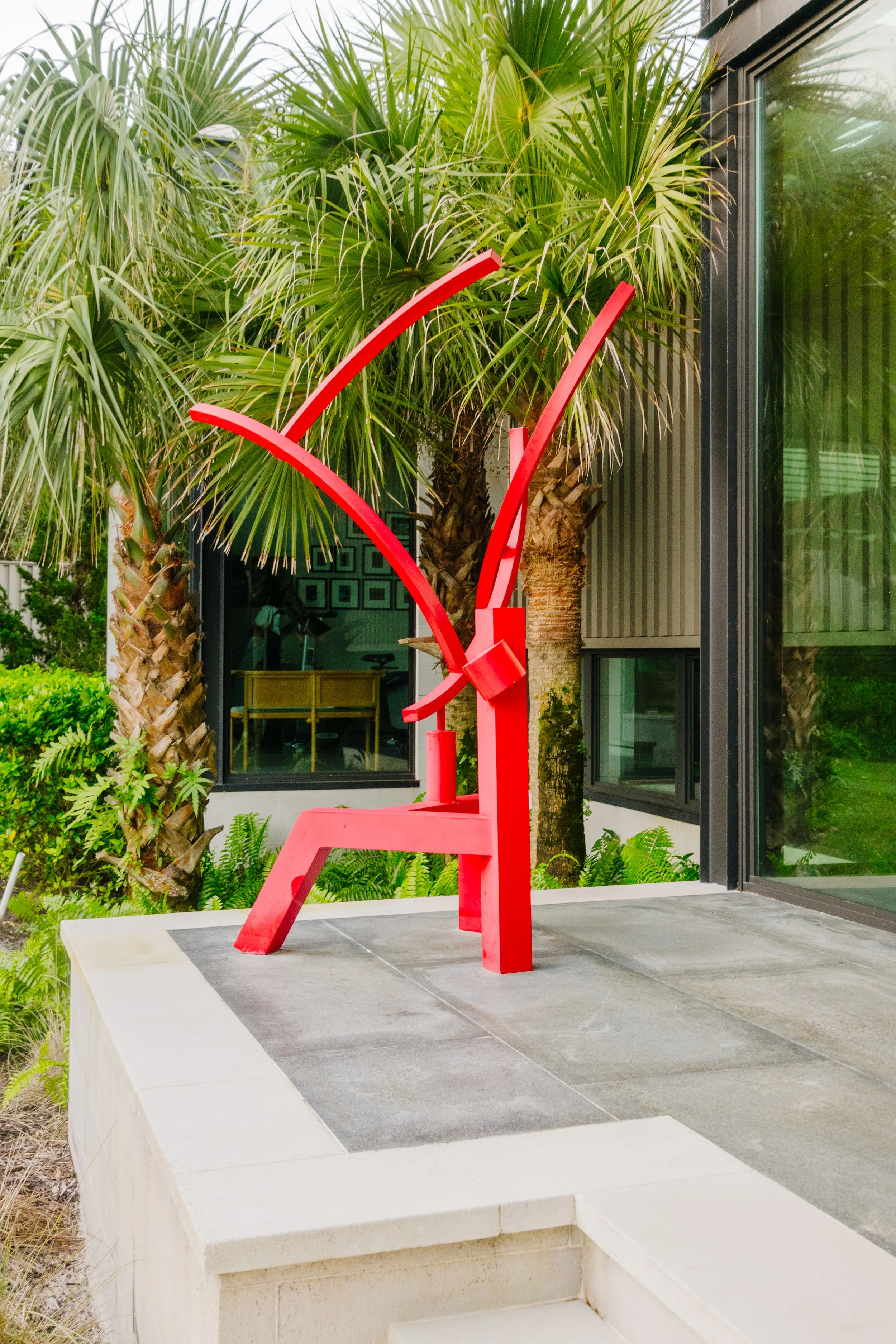 A red modern art sculpture on a concrete platform in an outdoor garden area with lush green palm trees and plants near a black framed glass building.