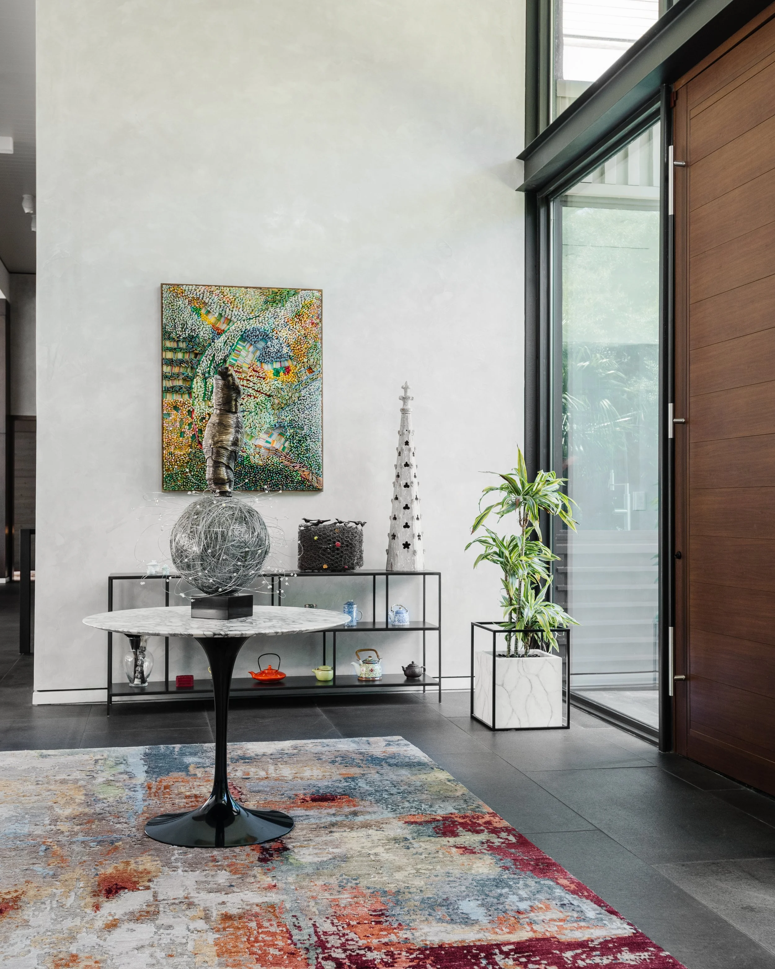 Modern interior with a round marble table, a colorful abstract painting on the wall, decorative sculptures, potted plants, and a sliding glass door allowing natural light.