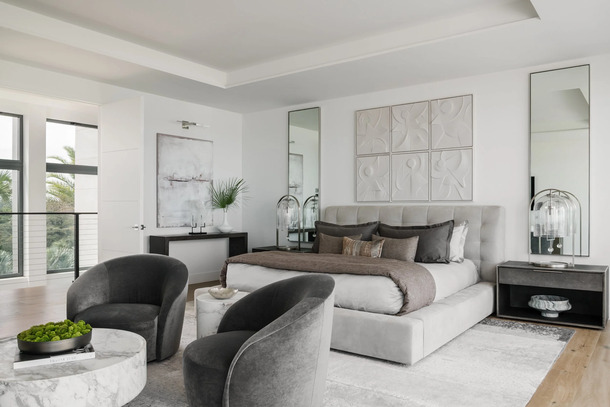 Modern living room with white walls, large windows, abstract wall art, mirrored panels, a beige tufted sofa with dark pillows, two glass table lamps, a black console table with a white vase and plant, and a marble coffee table with a green decorative