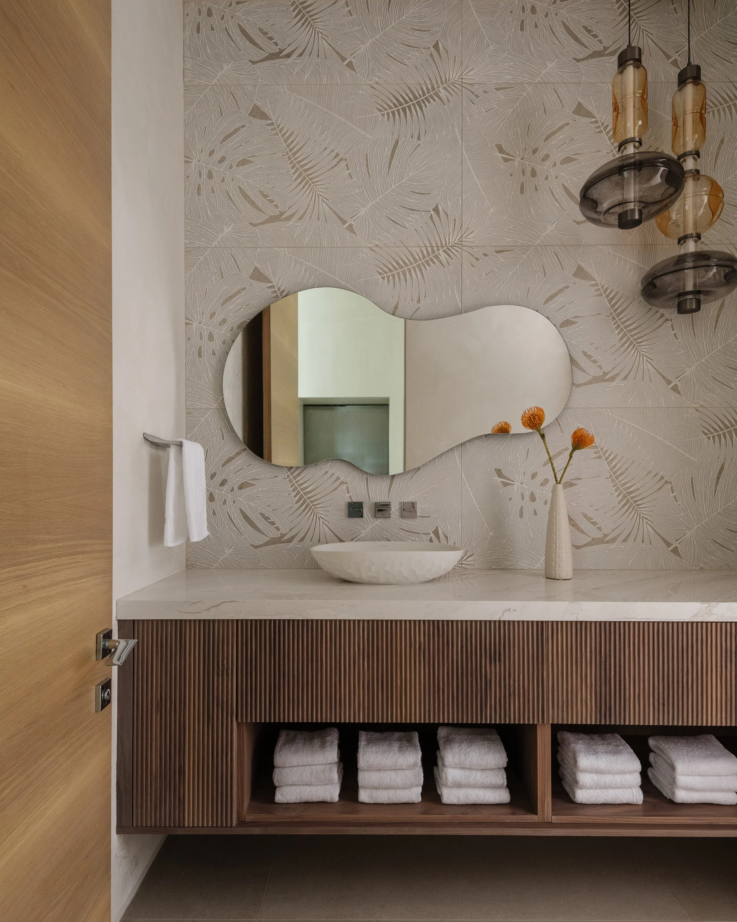 Modern bathroom vanity with a white marble countertop, a white vessel sink, a mirror with an abstract shape, a vase with orange flowers, a wall with leaf-patterned wallpaper, a hanging light fixture, and shelves with neatly rolled white towels.