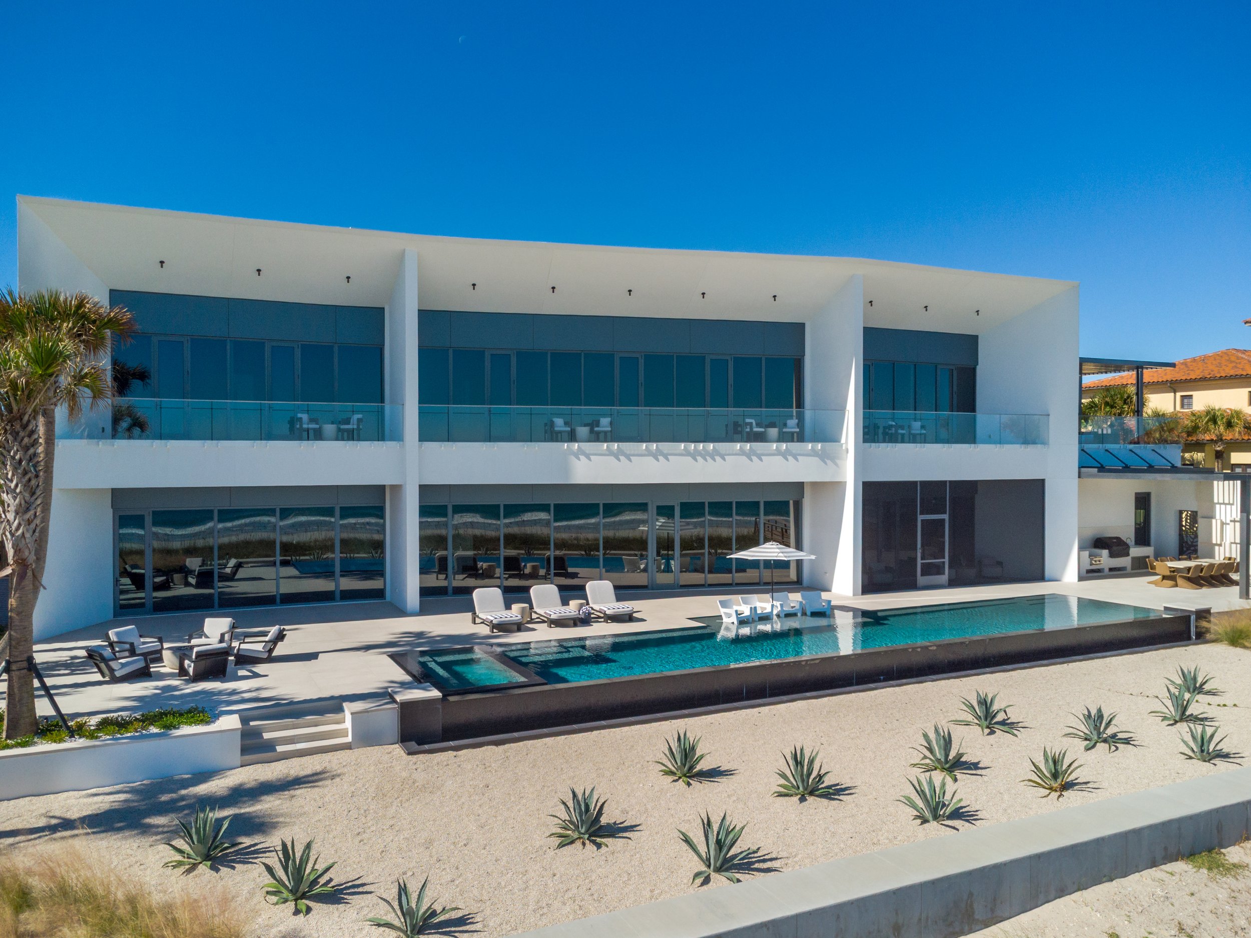 Modern two-story house with large glass windows, a balcony, outdoor seating area, and a swimming pool. Landscape includes desert plants and sandy ground, under a clear blue sky.