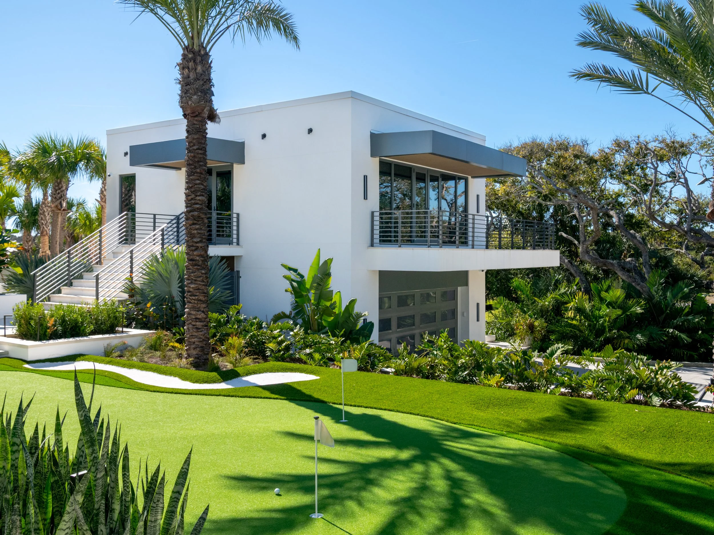 A modern white house with gray accents on a sunny day, surrounded by palm trees and lush greenery, with a well-maintained lawn and a putting green in the foreground.