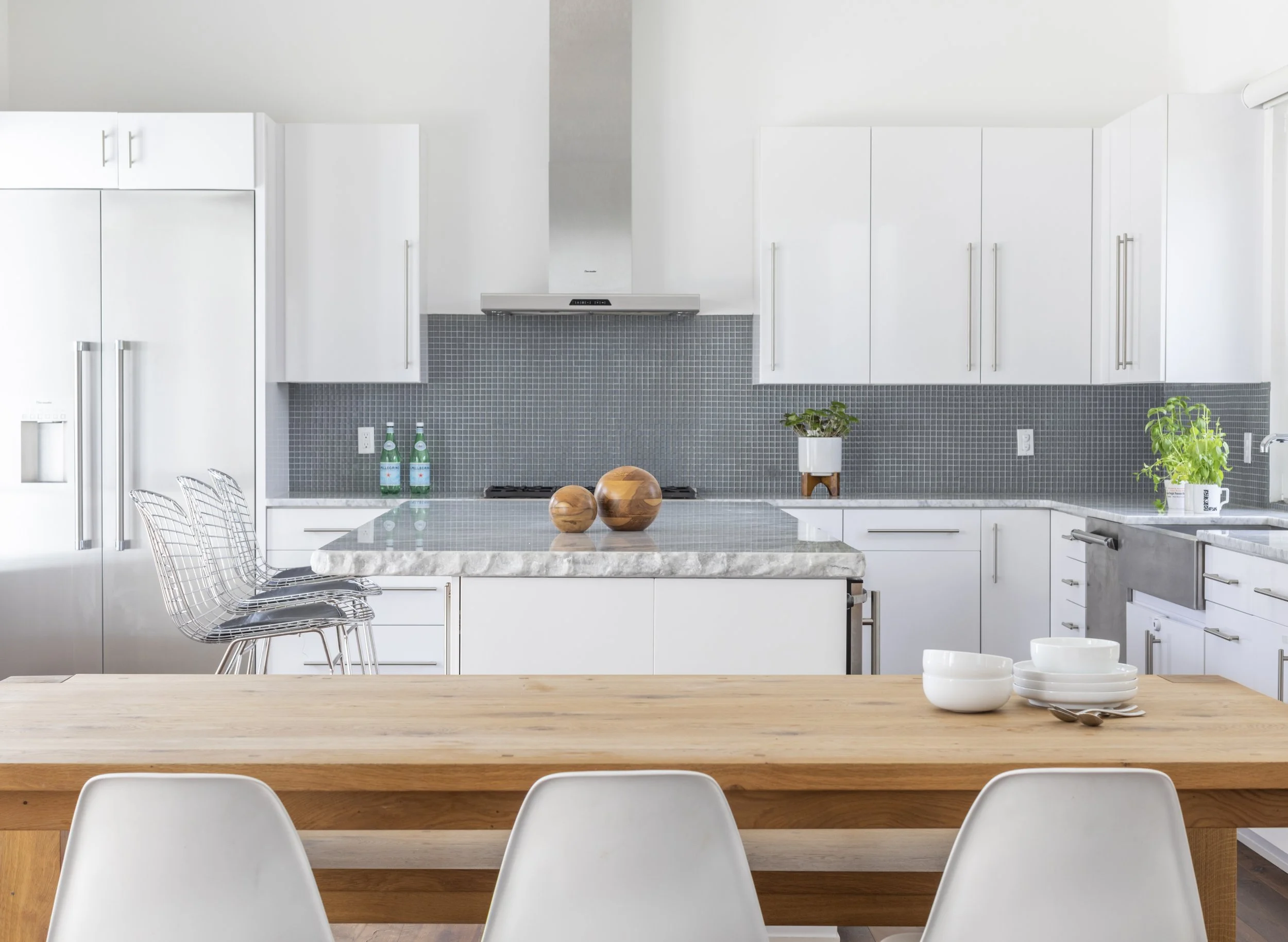 Modern kitchen with white cabinets, a gray tile backsplash, a white range hood, a marble kitchen island with wooden accents, white bowls, and a dining table with white chairs and a wooden top.