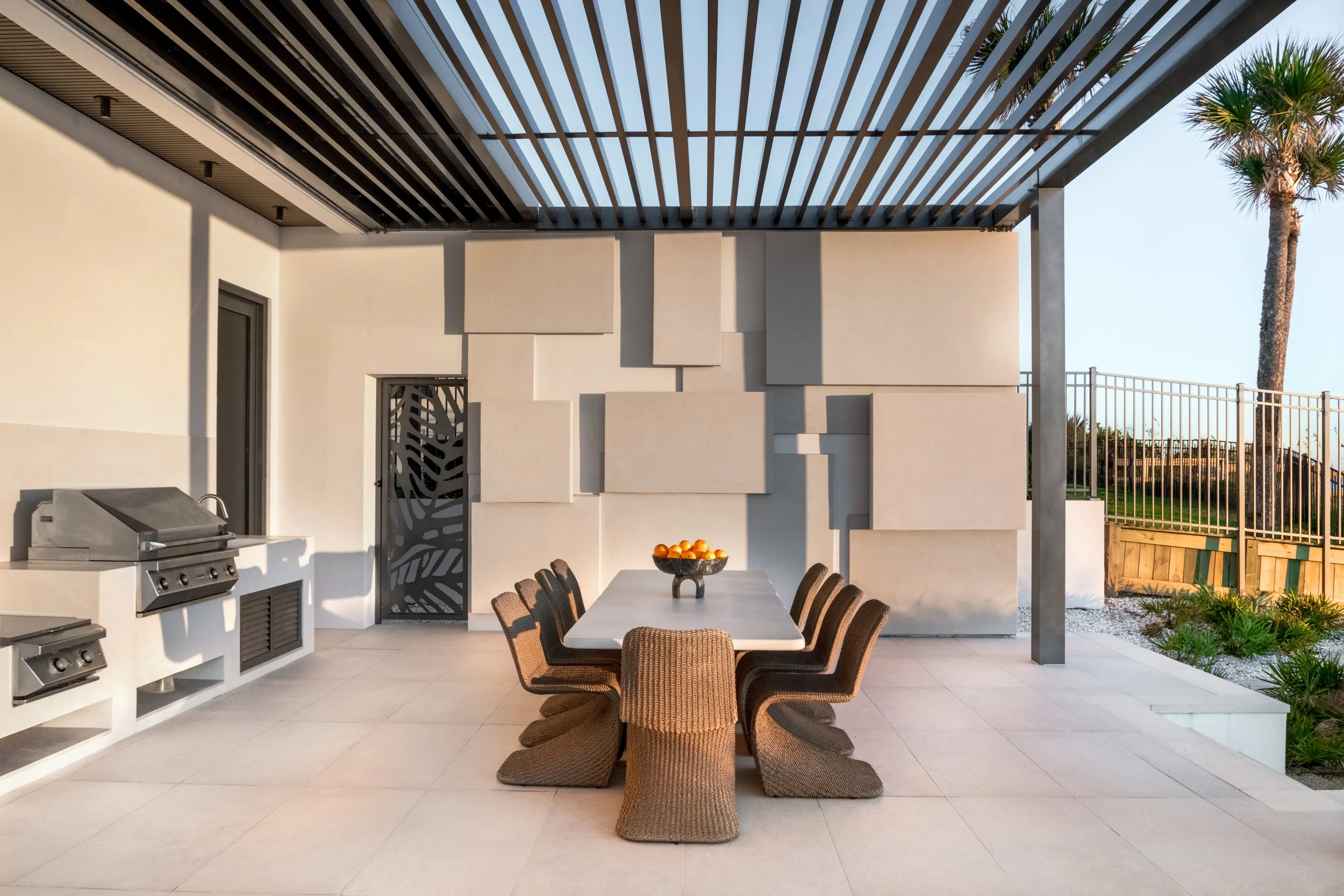 Outdoor patio area with a dining table and wicker chairs, a wall with decorative panels, a grill, and a view of palm trees and a fence in the background.