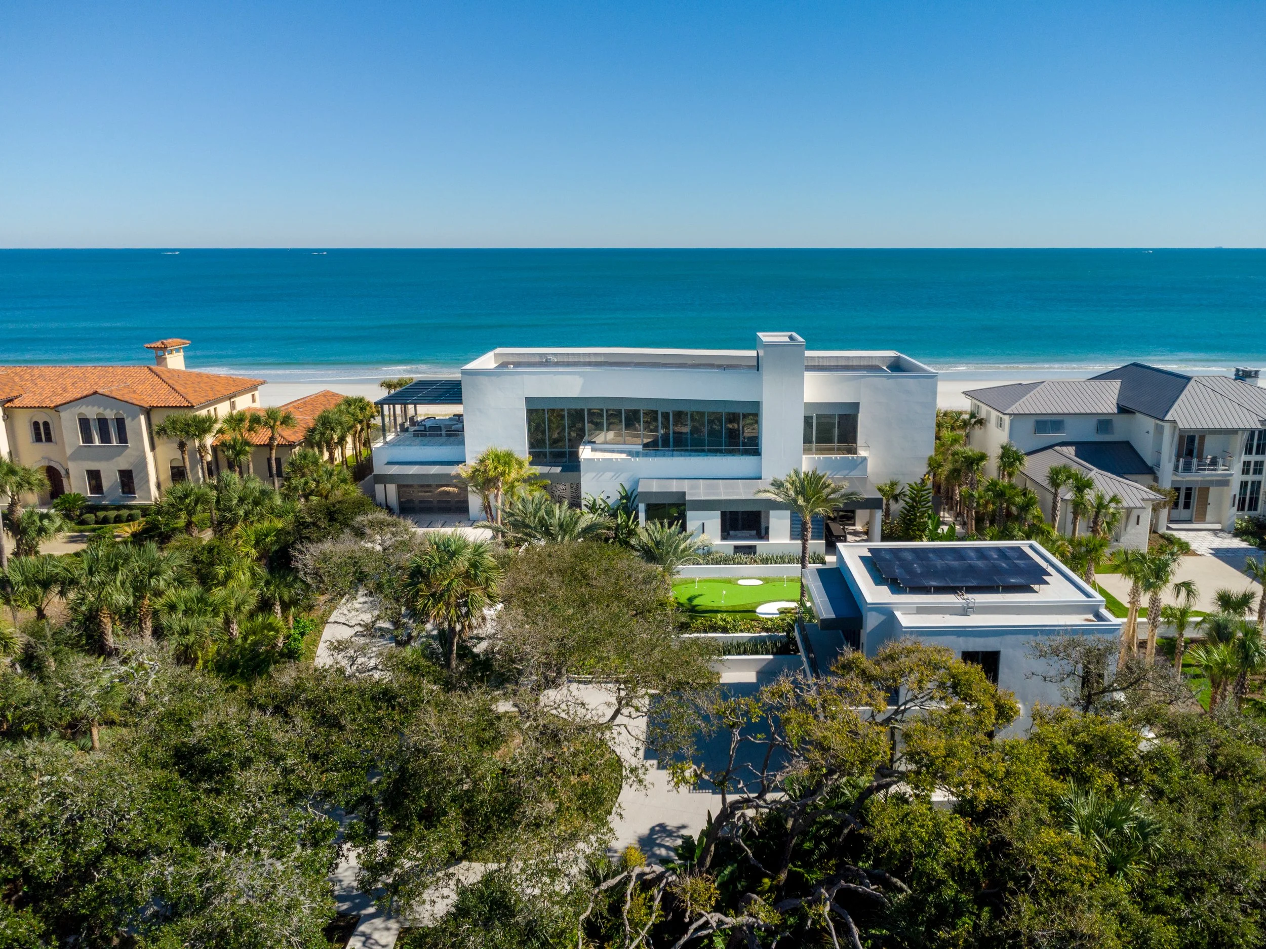 Aerial view of a coastal neighborhood with modern white houses, lush greenery, and the ocean in the background under a clear blue sky.