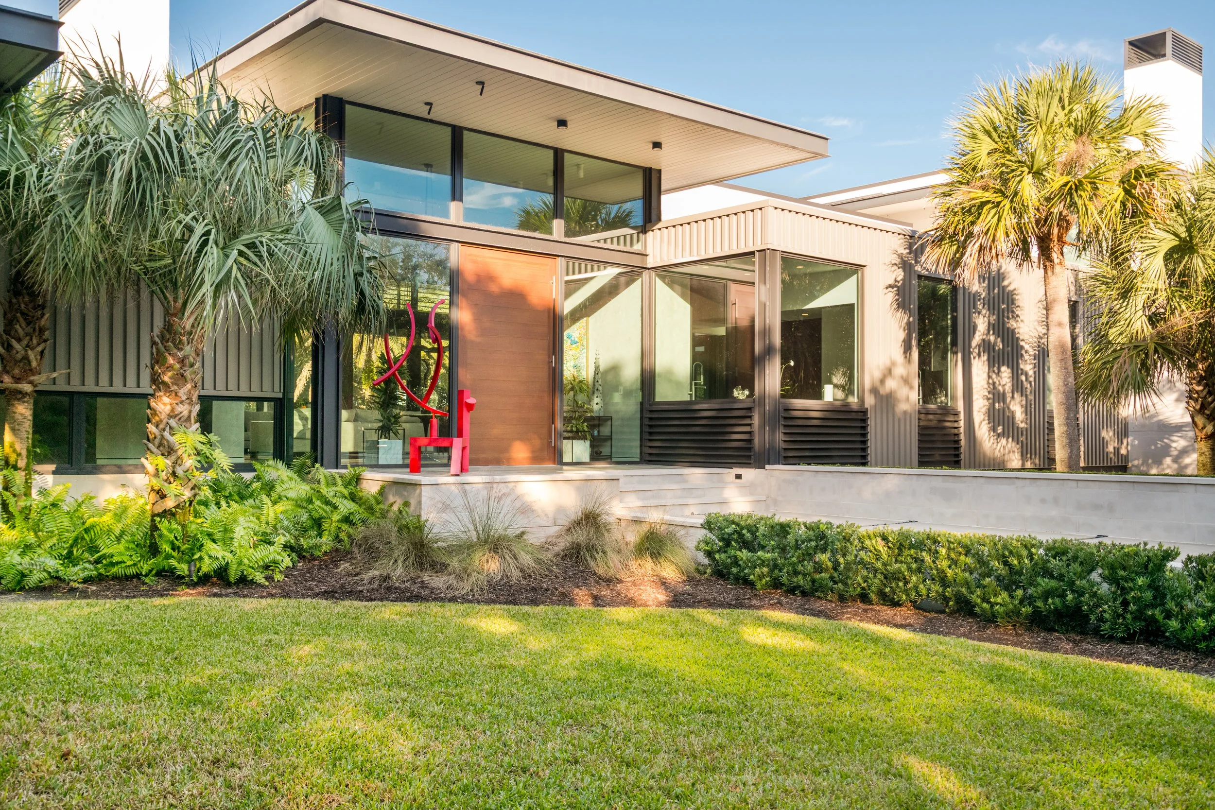 Modern house with large glass windows, surrounded by lush greenery including palm trees and shrubs, with a well-maintained lawn in the foreground.