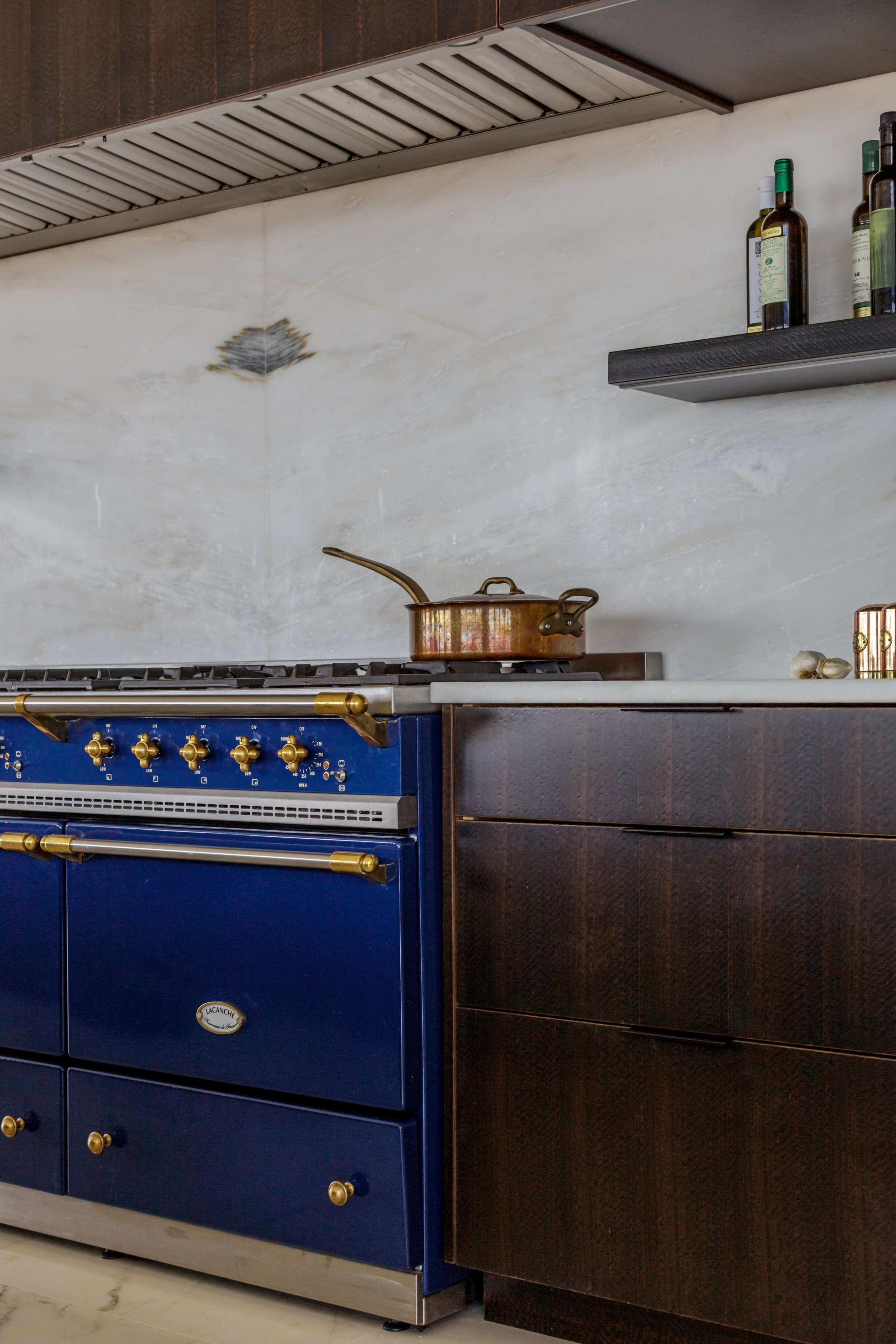 A kitchen scene showing a gas stove with a copper pot on top, a blue oven with gold knobs, and a black shelf holding bottles of olive oil. The background features a beige marble wall with a dark wood cabinet above.