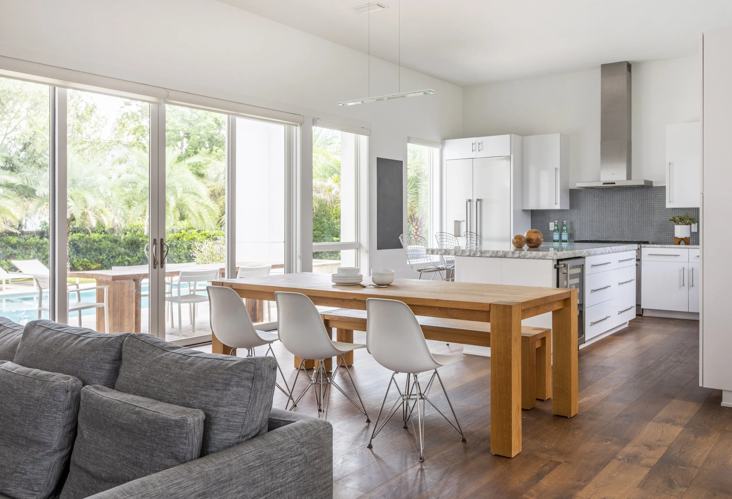 Modern open-concept kitchen and dining area with large sliding glass doors leading to an outdoor patio and pool, white cabinets, marble countertops, wooden dining table with white chairs, gray couch in the foreground, wooden flooring.