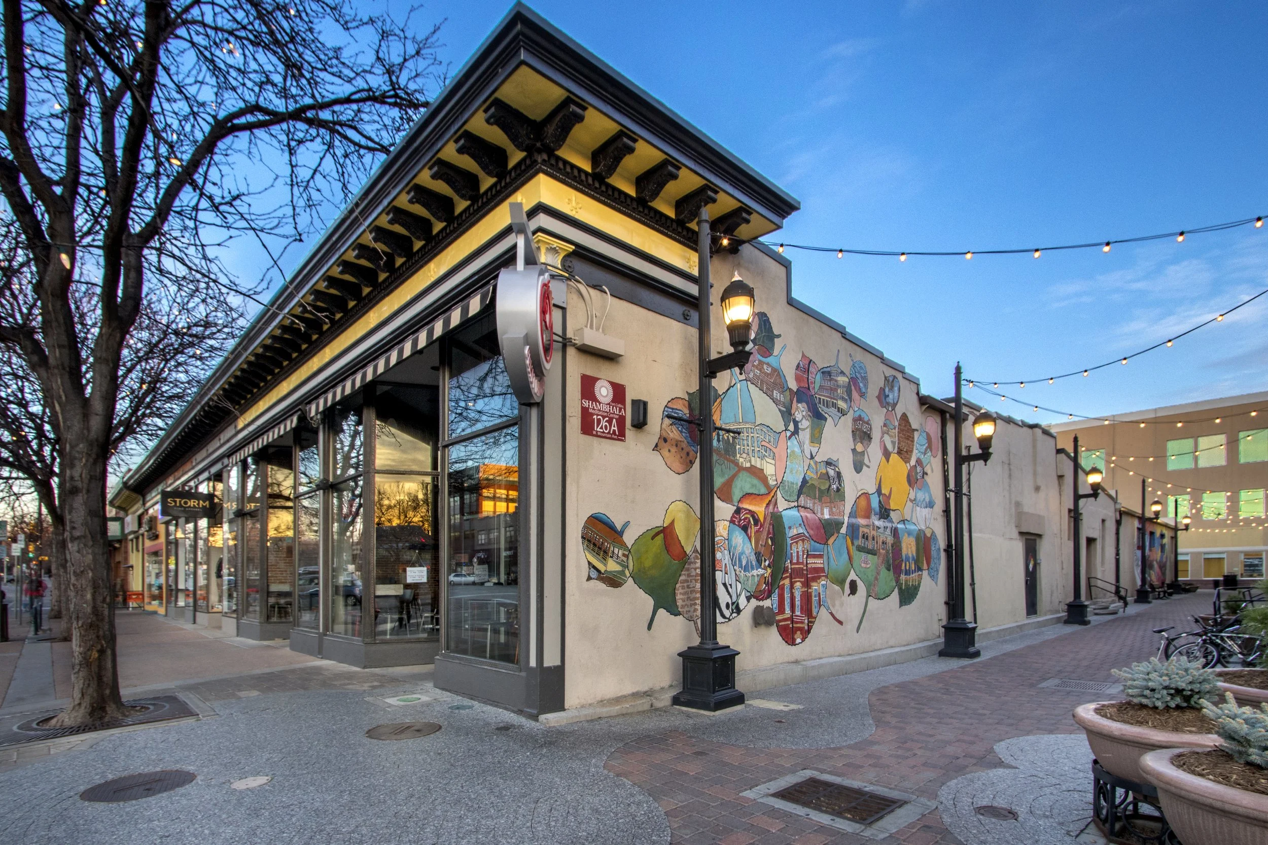 An outdoor urban scene at dusk featuring a corner building with a mural of the United States map, decorated with local landmarks and symbols. The building has glass storefront windows, black trim, and a yellow awning. There are street lamps and tree branches in the foreground, with string lights overhead, and neighboring buildings with large windows.