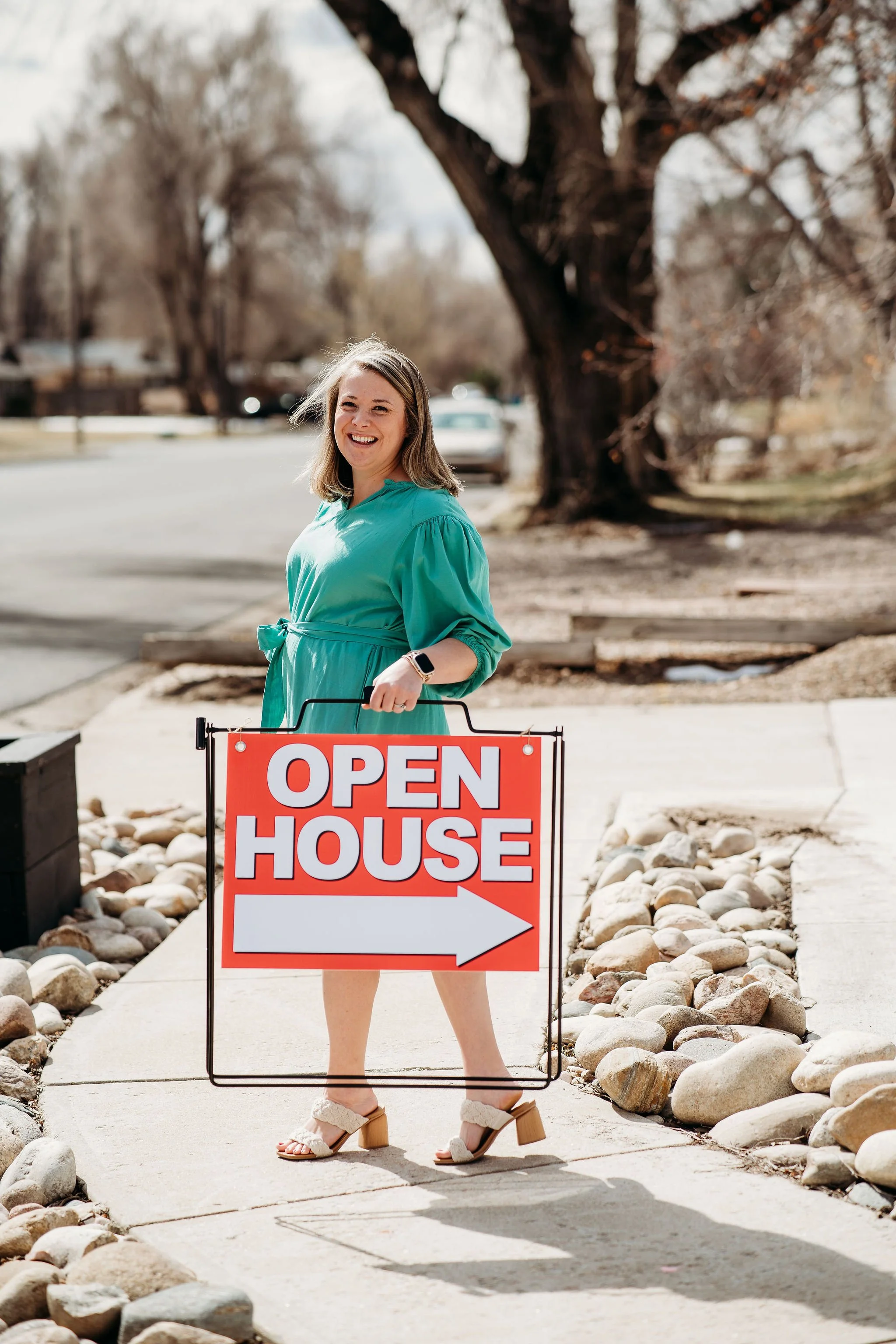 Woman standing outdoors holding a sign that reads "Open House" with an arrow pointing to the right, smiling, wearing a turquoise dress and tan heeled sandals, with large rocks lining the sidewalk and a large tree in the background.